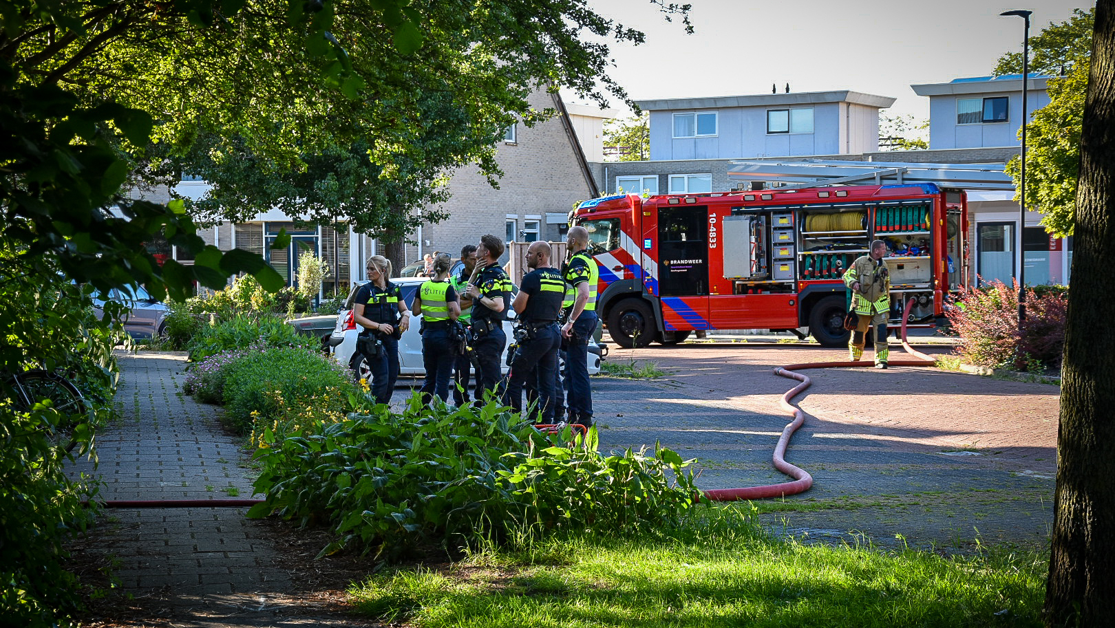 Politieagenten praten samen voor een geparkeerde brandweerwagen, met een brandweerman die een brandslang vasthoudt.