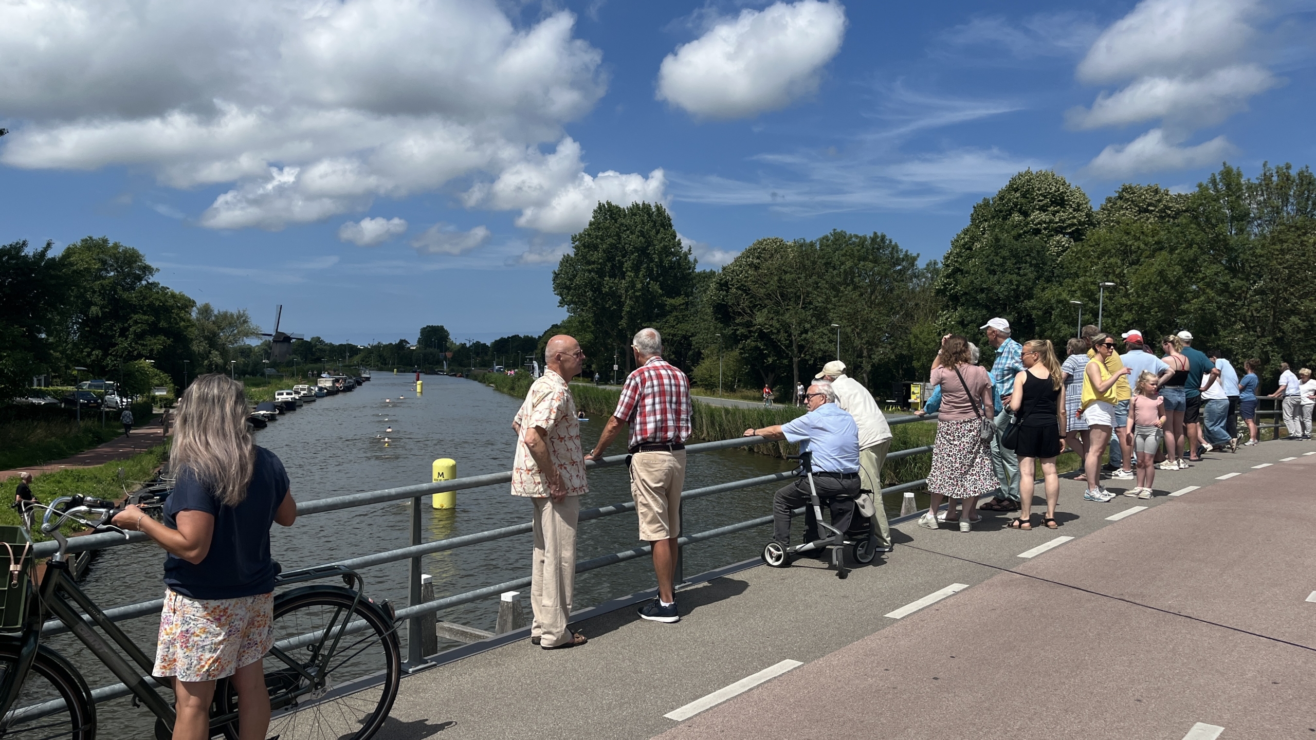 Mensen staan op een brug langs een rivier en kijken naar iets in de verte; op de achtergrond is een windmolen te zien onder een deels bewolkte lucht.