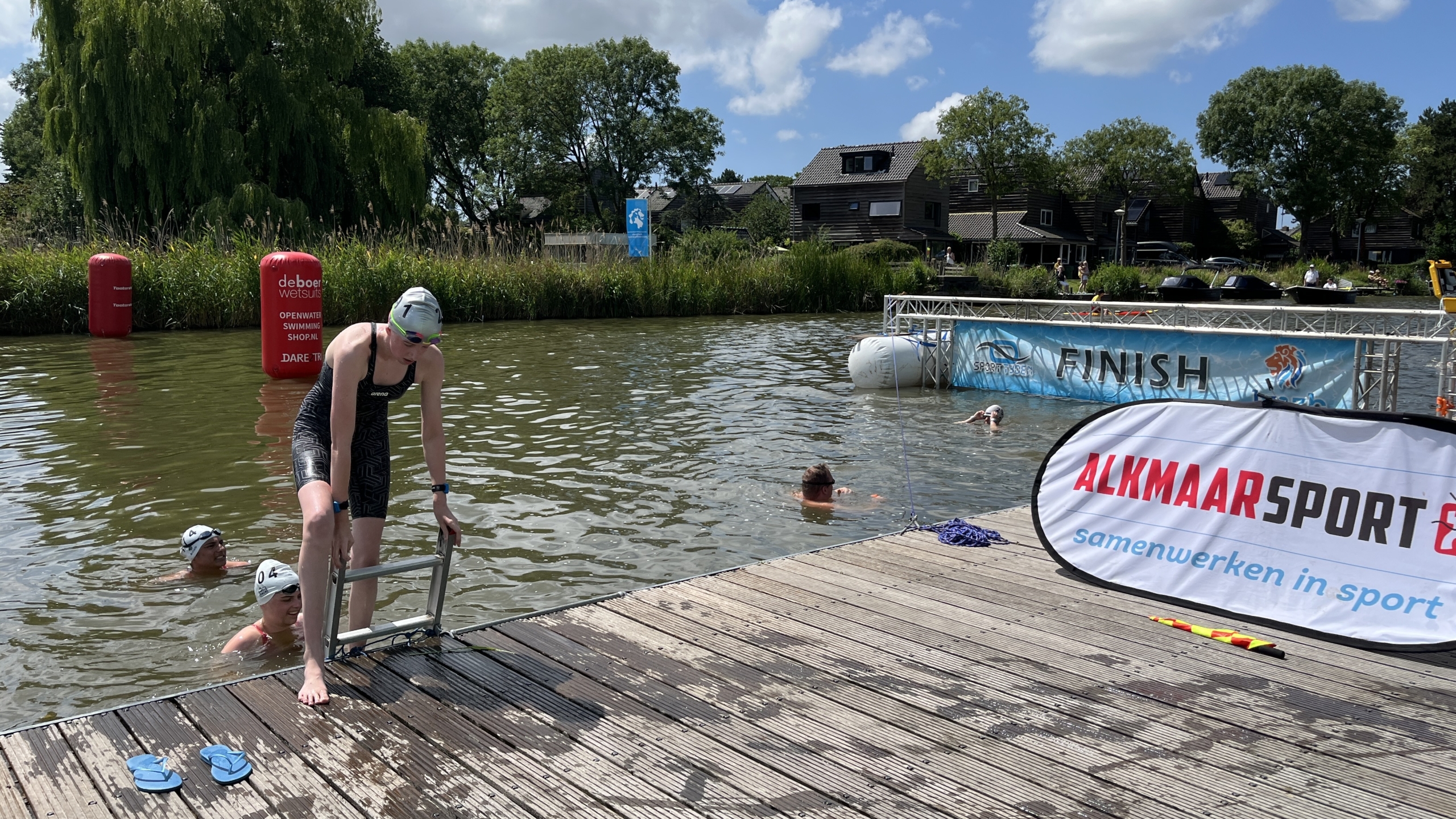 Een zwemmer komt uit het water op een steiger tijdens een wedstrijd, omringd door andere zwemmers. Op de achtergrond zijn finishvlaggen en het merk "ALKMAARSPORT" te zien.