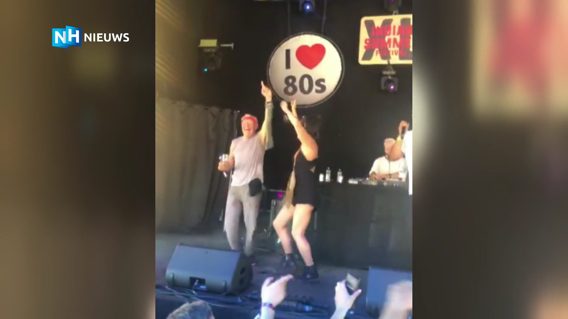 Twee mensen dansen op een podium met een bord "I Love 80s" op de achtergrond en een DJ op de achtergrond.