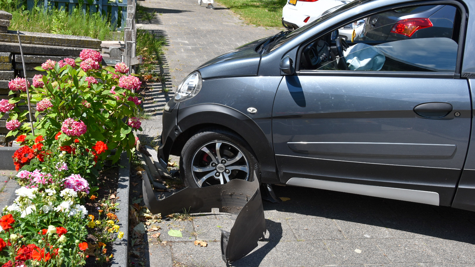 Een grijze auto is tegen een bloemperk gebotst, met bloemen zoals hortensia's en andere kleurrijke bloemen, en de voorbumper is beschadigd.