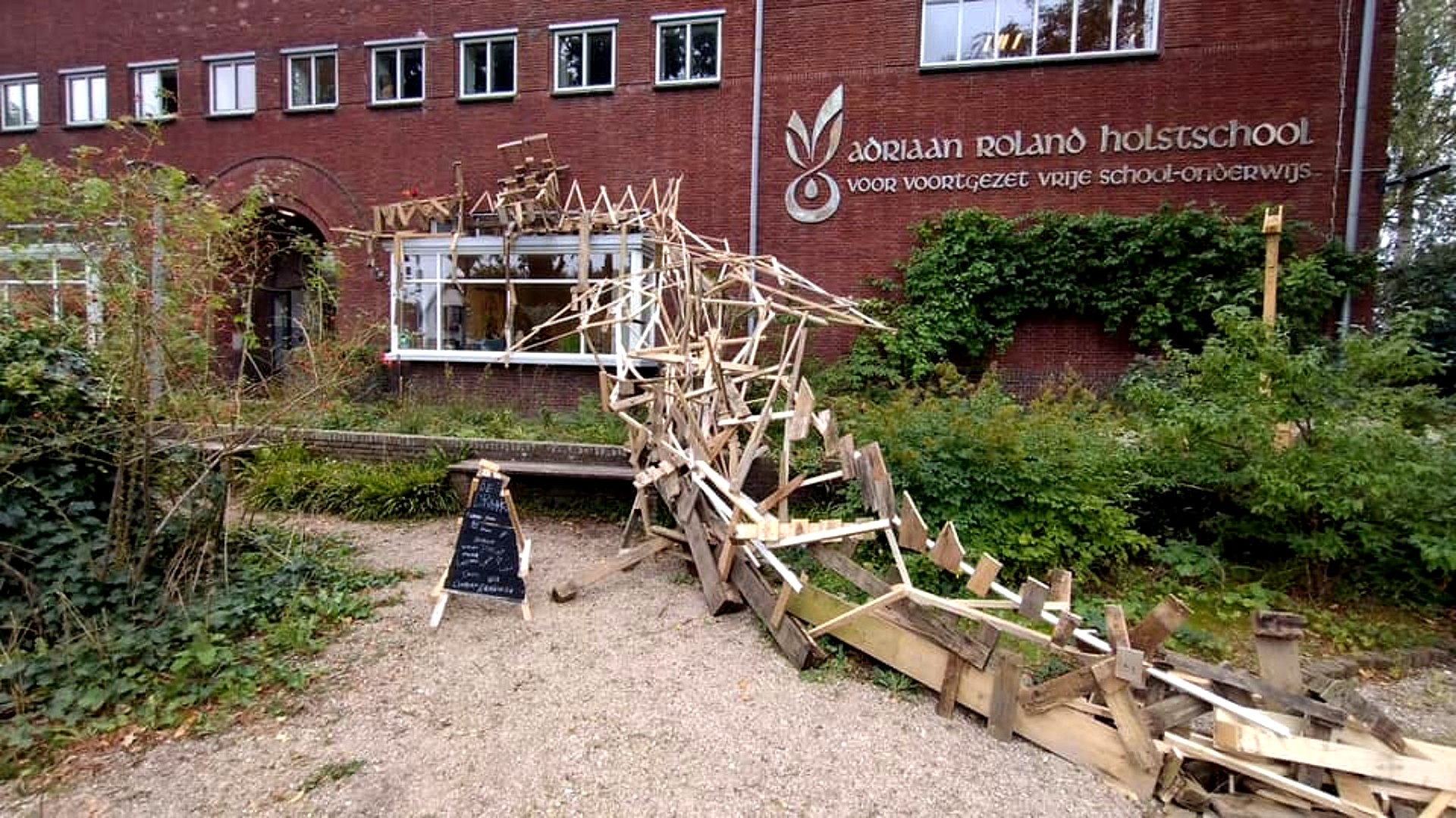 Buiten kunstinstallatie van houten planken in organische vormen voor de Adriaan Roland Holstschool, met groene begroeiing en een schoolbord ernaast.