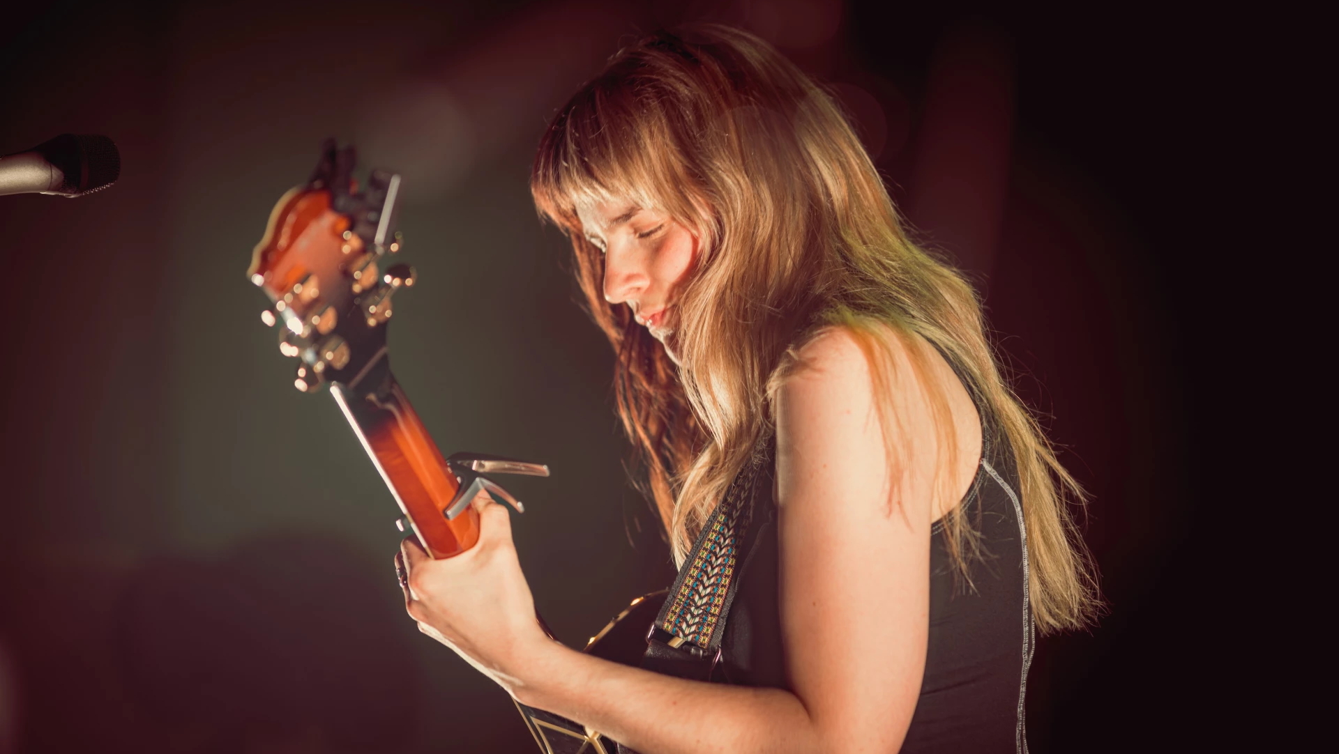 Vrouw met lang haar speelt gitaar op een podium, gehuld in sfeervol licht.