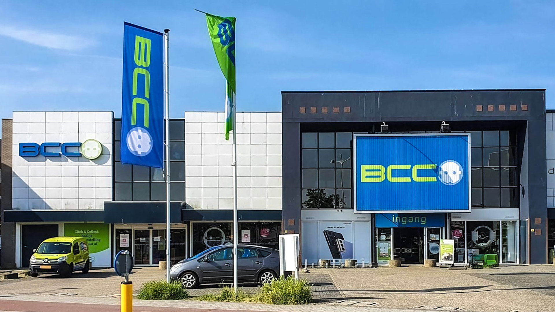 Voorzijde van een BCC-winkel met blauwe en groene reclameborden, een parkeerplaats met enkele auto's en vlaggen met het BCC-logo.