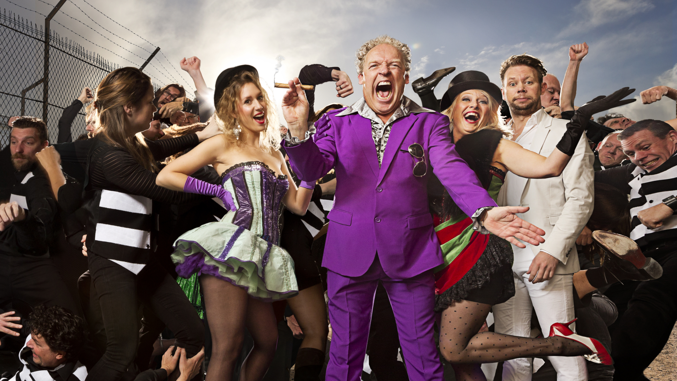 Een kleurrijke groep mensen in extravagante kleding, waaronder een man in een paars pak en vrouwen in kostuums, staan te midden van een rumoerige menigte voor een hek met prikkeldraad.