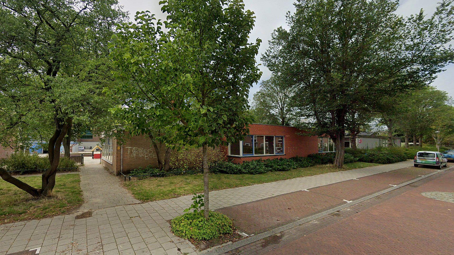 Straatbeeld met een laag gebouw met rode bakstenen en ramen, omgeven door bomen en een tuin; een trottoir en geparkeerde auto zijn zichtbaar op de voorgrond.