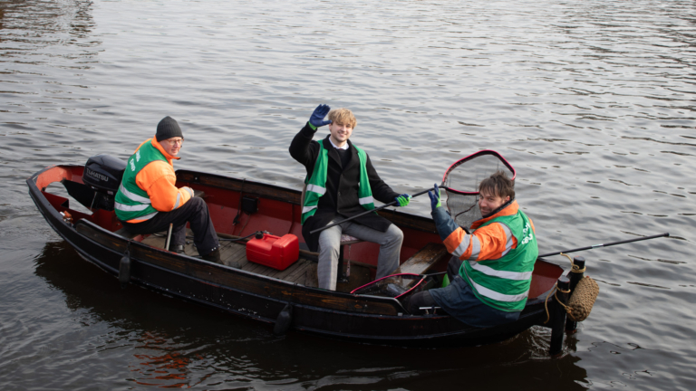 Drie mensen in een kleine boot op een rivier, met een motor en een rode brandstoftank. Ze dragen groene veiligheidshesjes en houden een schepnet vast; één van hen zwaait naar de camera.