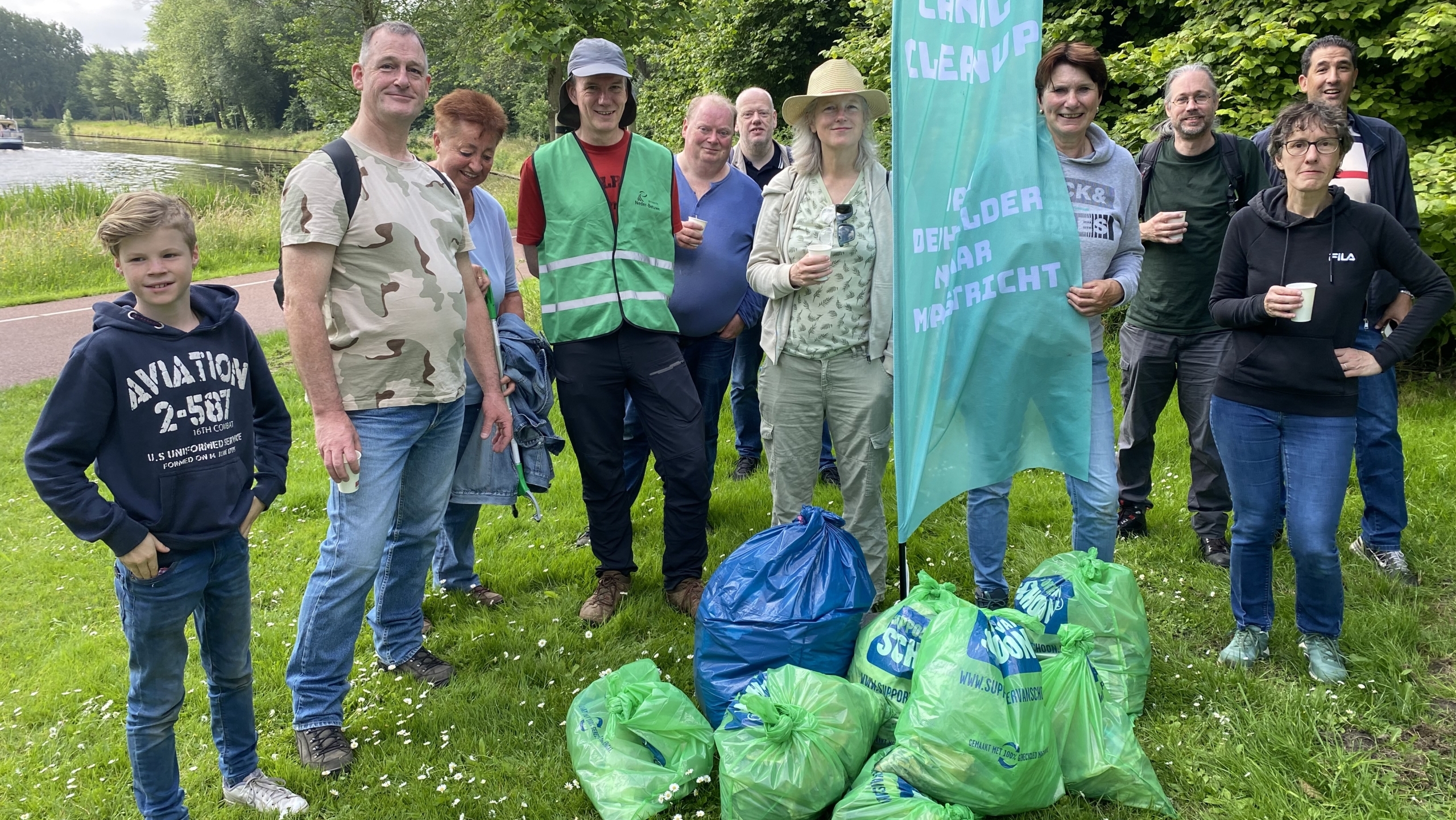 Een groep mensen staat bij elkaar in een parkachtige omgeving met afvalzakken vol zwerfvuil voor hen, naast een vlag met de tekst 'Canal Cleanup'.
