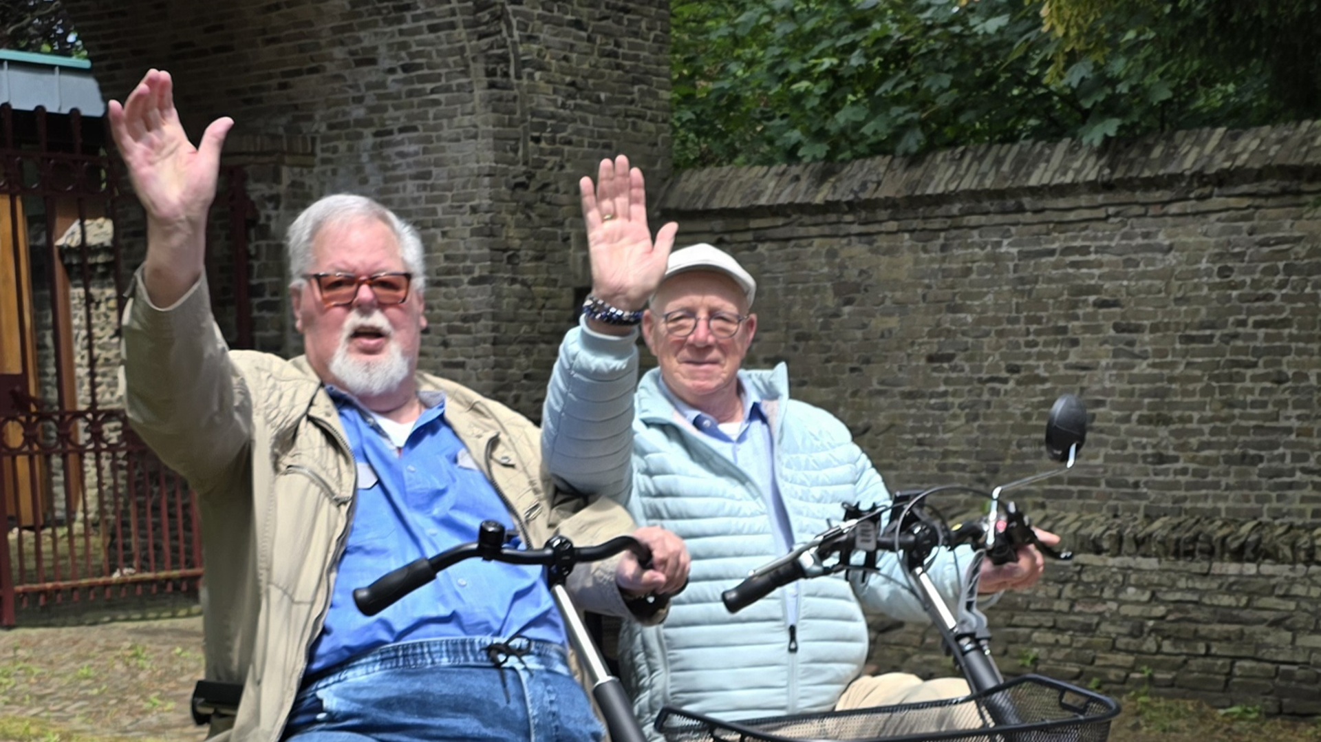 Twee oudere mannen zwaaien terwijl ze op een scooter rijden tegen een achtergrond van oude stenen muren en groen.