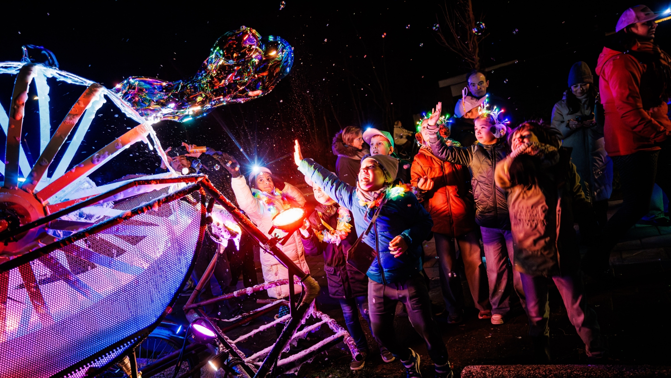 Een groep mensen, waaronder kinderen, staat 's nachts in felle verlichting met hoofdlampen en kleurrijke kleding terwijl ze kijken naar een grote zeepbel die door een verlichte installatie wordt geblazen.