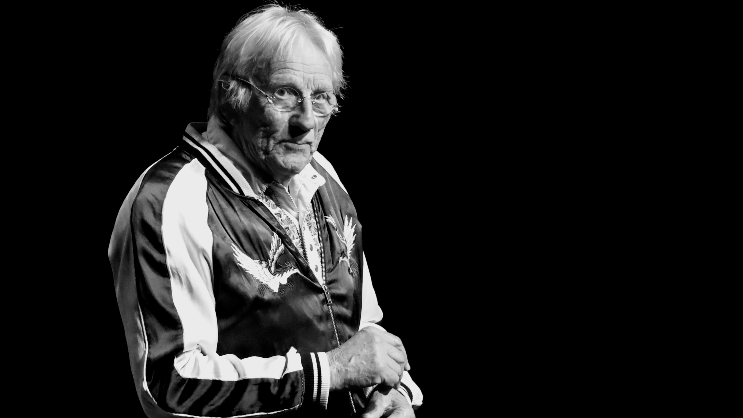 Oudere man met wit haar en bril, gekleed in een jas met adelaarmotief, staat op een podium in zwart-witfoto.