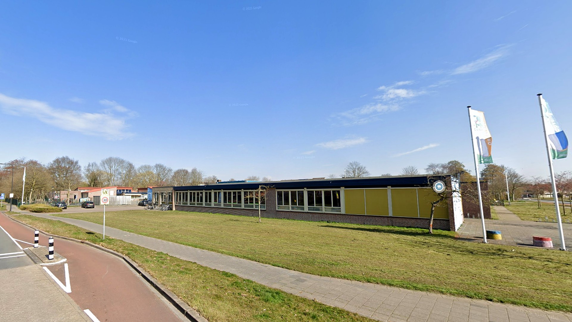 Lang, laag gebouw met veel ramen aan de rechterkant van de afbeelding; grasveld en trottoir op de voorgrond; vlaggen en een verkeersbord op de achtergrond; heldere blauwe lucht.