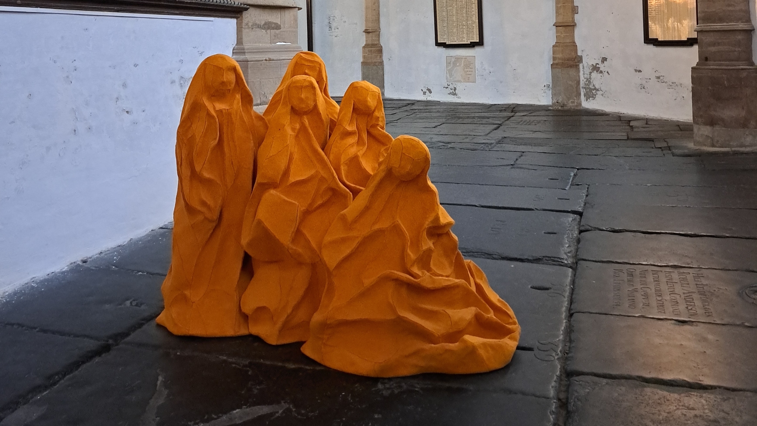 Oranje sculptuur van drie figuren in gewaden, geplaatst in een historische kerk met stenen vloer en gebrandschilderde ramen.