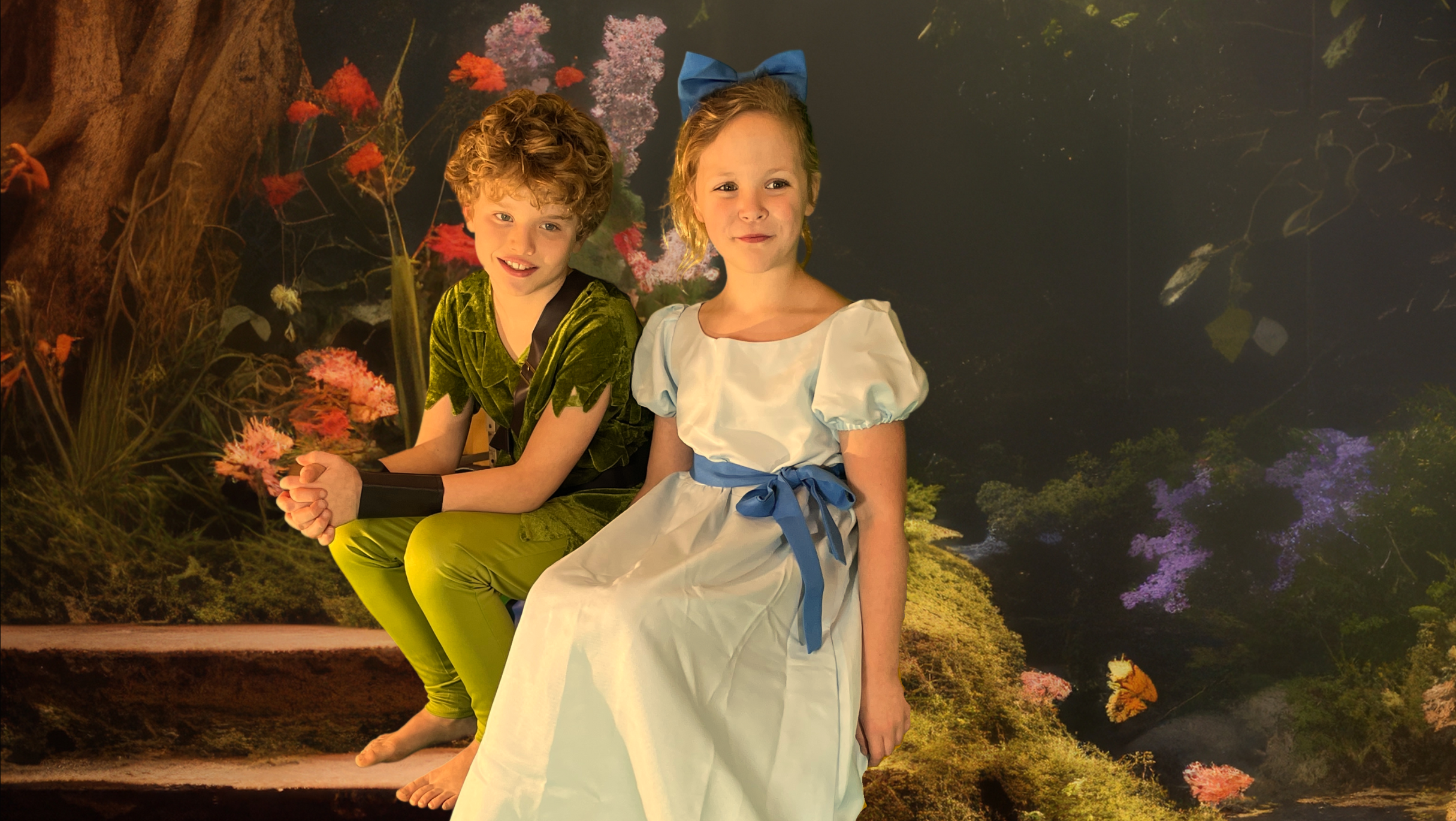 Twee kinderen verkleed als sprookjesfiguren, zittend in een bosachtige omgeving met kleurrijke bloemen; het meisje draagt een lichtblauwe jurk met pofmouwen en een strik, en de jongen draagt een groen kostuum met een riem en leggings.