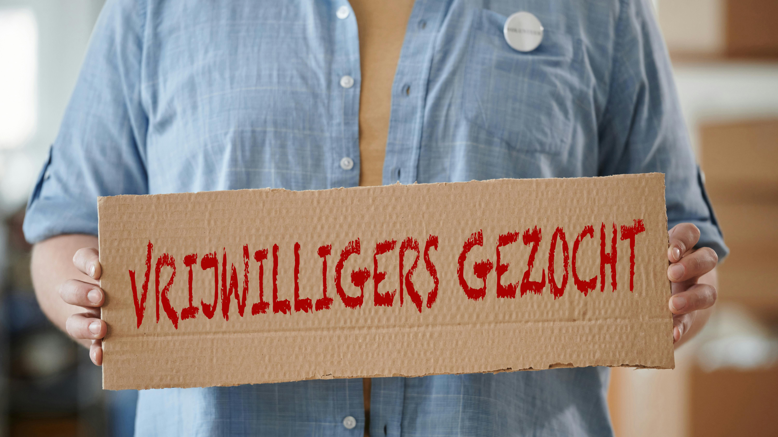 Een persoon houdt een kartonnen bord vast met daarop de tekst 'VRIJWILLIGERS GEZOCHT' in rode letters.