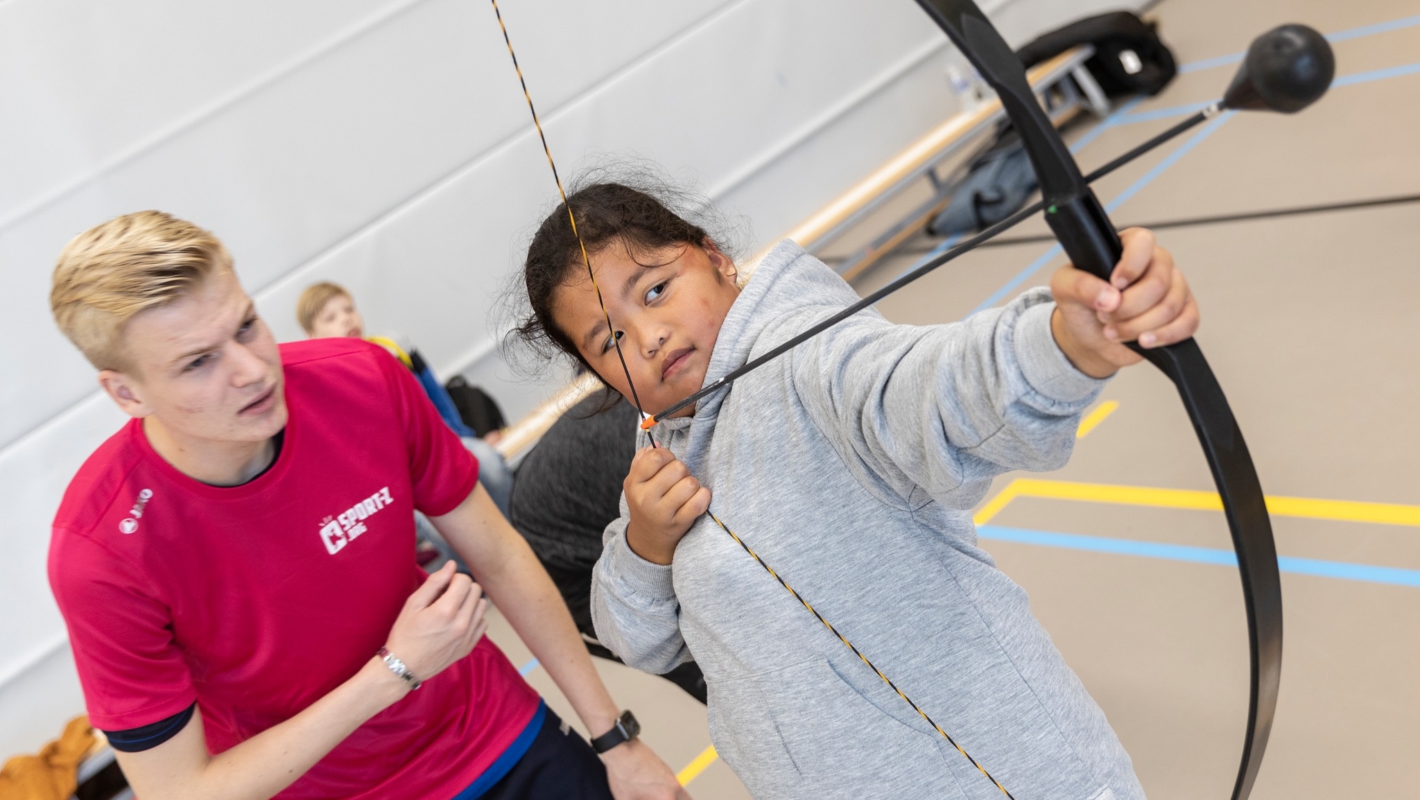 Een kind in een grijze hoodie richt een boog en pijl, terwijl een jonge man in een rood sportshirt assisteert, binnen een gymzaal.