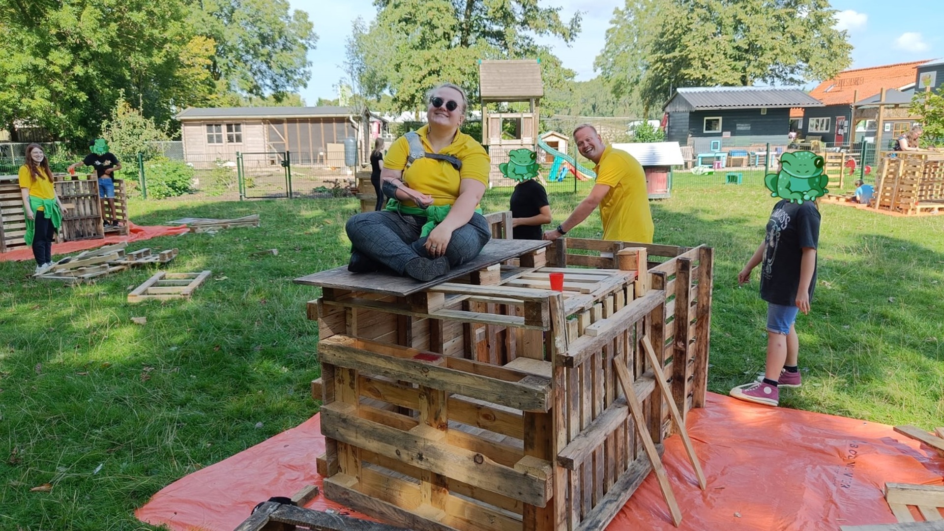 Mensen bouwen een structuur van houten pallets in een buitengebied, met een vrouw die op de constructie zit; enkele gezichten zijn bedekt met cartoonkikkers.