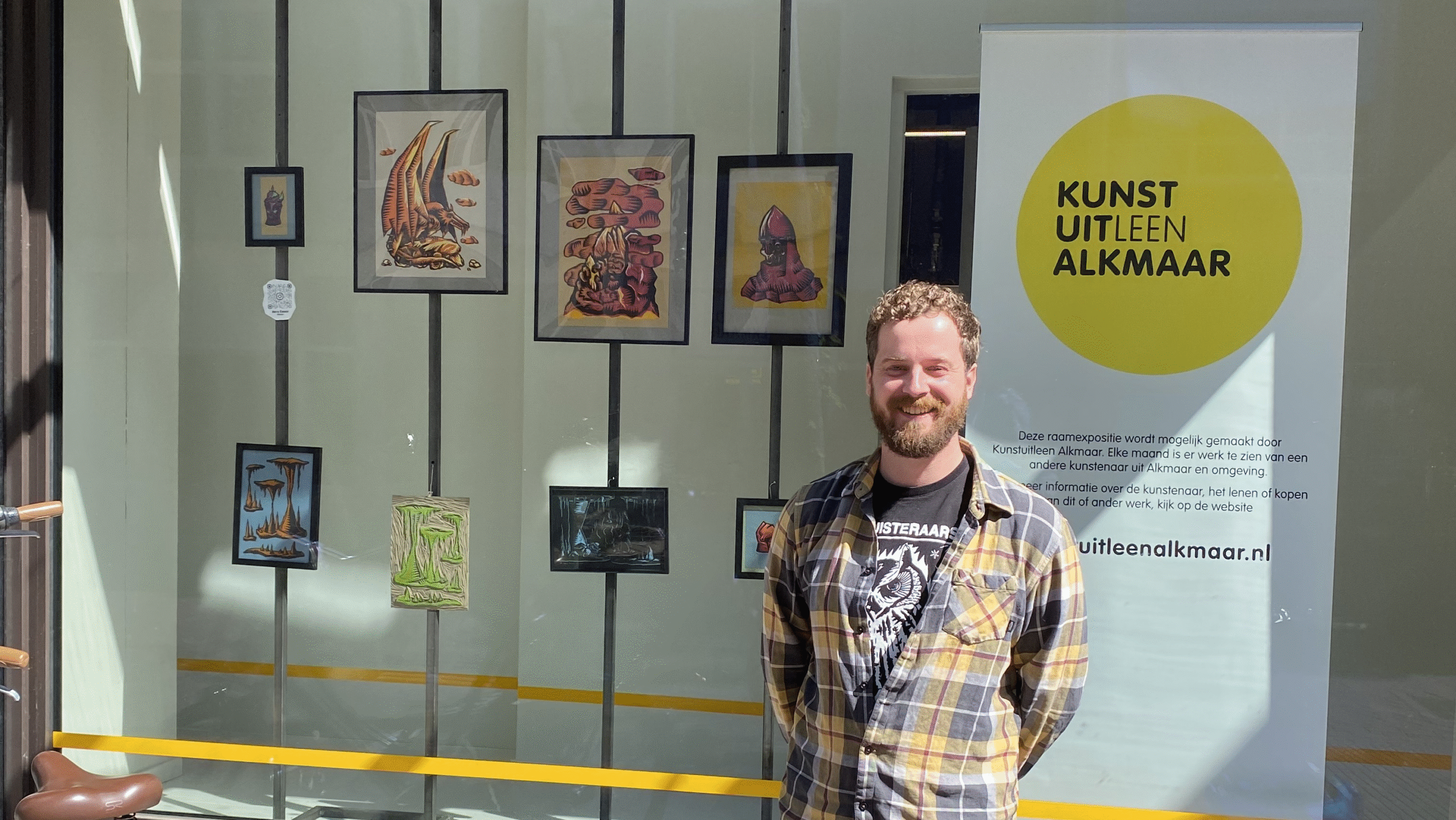 Man in checkered shirt smiling in front of a window display with artwork and a sign reading 'Kunstuitleen Alkmaar.'
