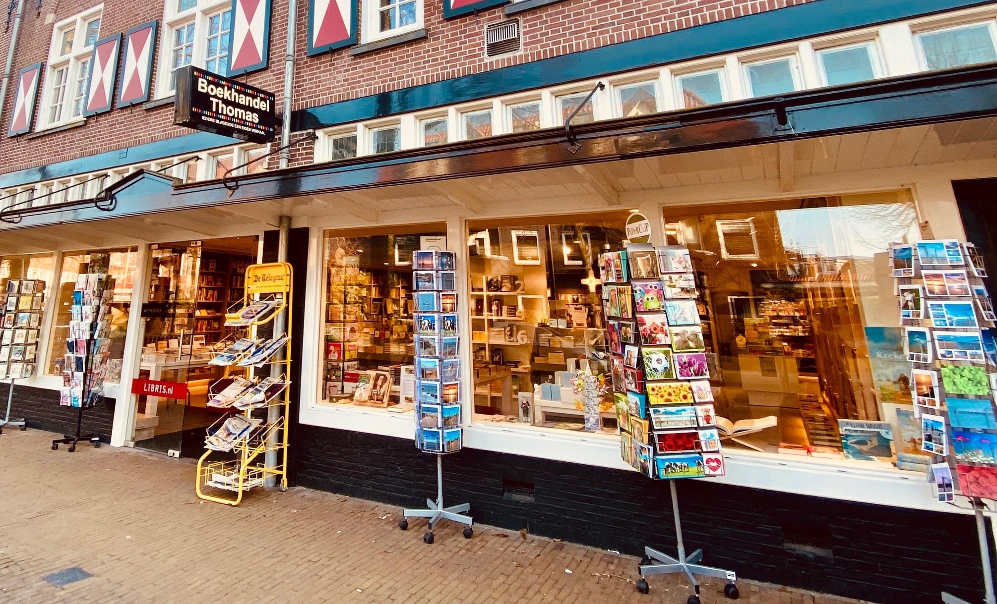 Buitenkant van een boekwinkel genaamd 'Boekhandel Thomas' met verschillende stellingen vol ansichtkaarten voor de etalage.