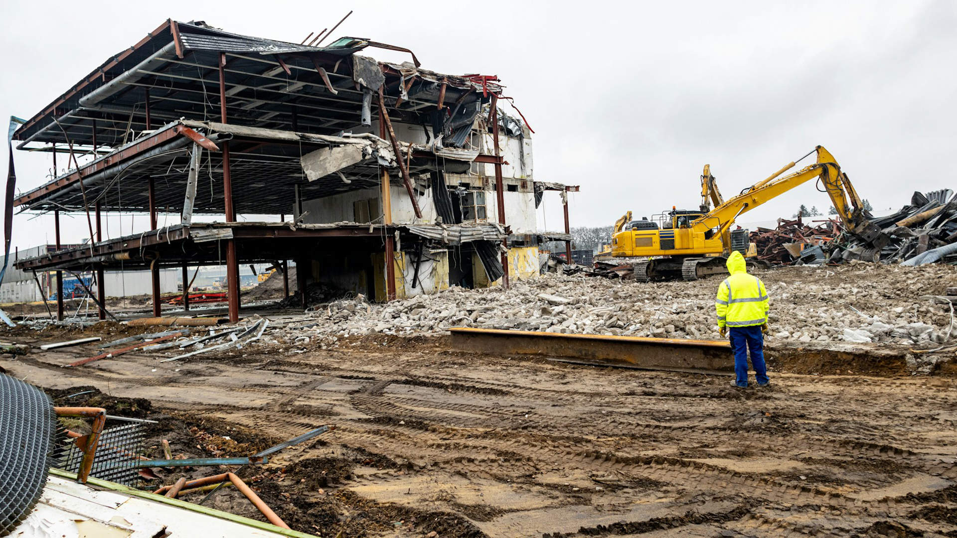 Een bouwvakker in een gele jas kijkt naar een sloopterrein met een half afgebroken gebouw en een gele graafmachine.