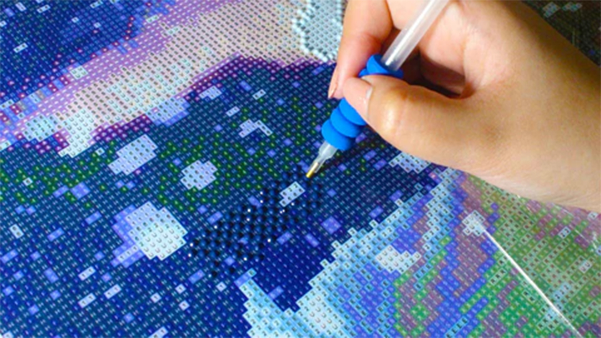 Een hand plaatst blauwe steentjes op een diamond painting canvas met een speciale pen, te midden van een patroon van blauwe en paarse vakjes.