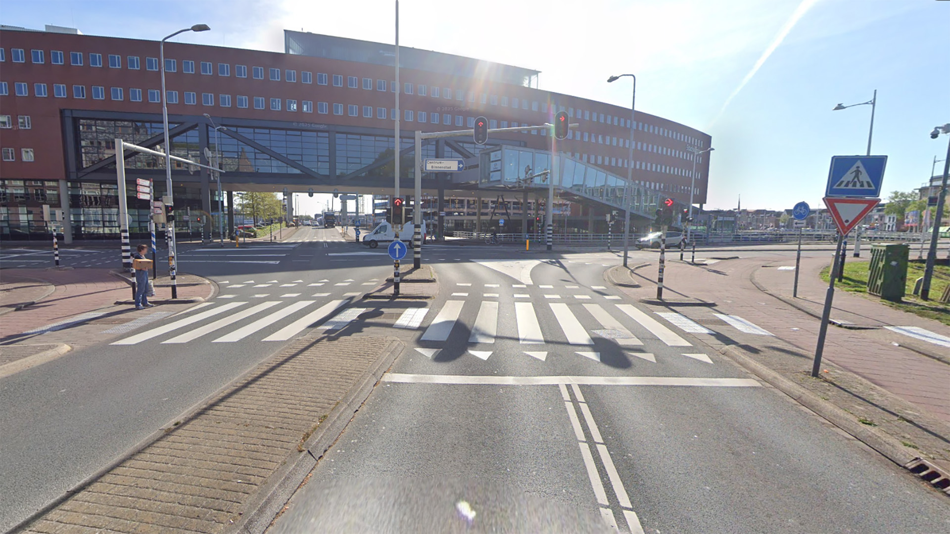 Straathoek met verkeerslichten en zebrapaden, met een groot, modern gebouw op de achtergrond dat over de weg hangt. Er zijn diverse verkeersborden zichtbaar en een voetganger wacht bij een zebrapad.