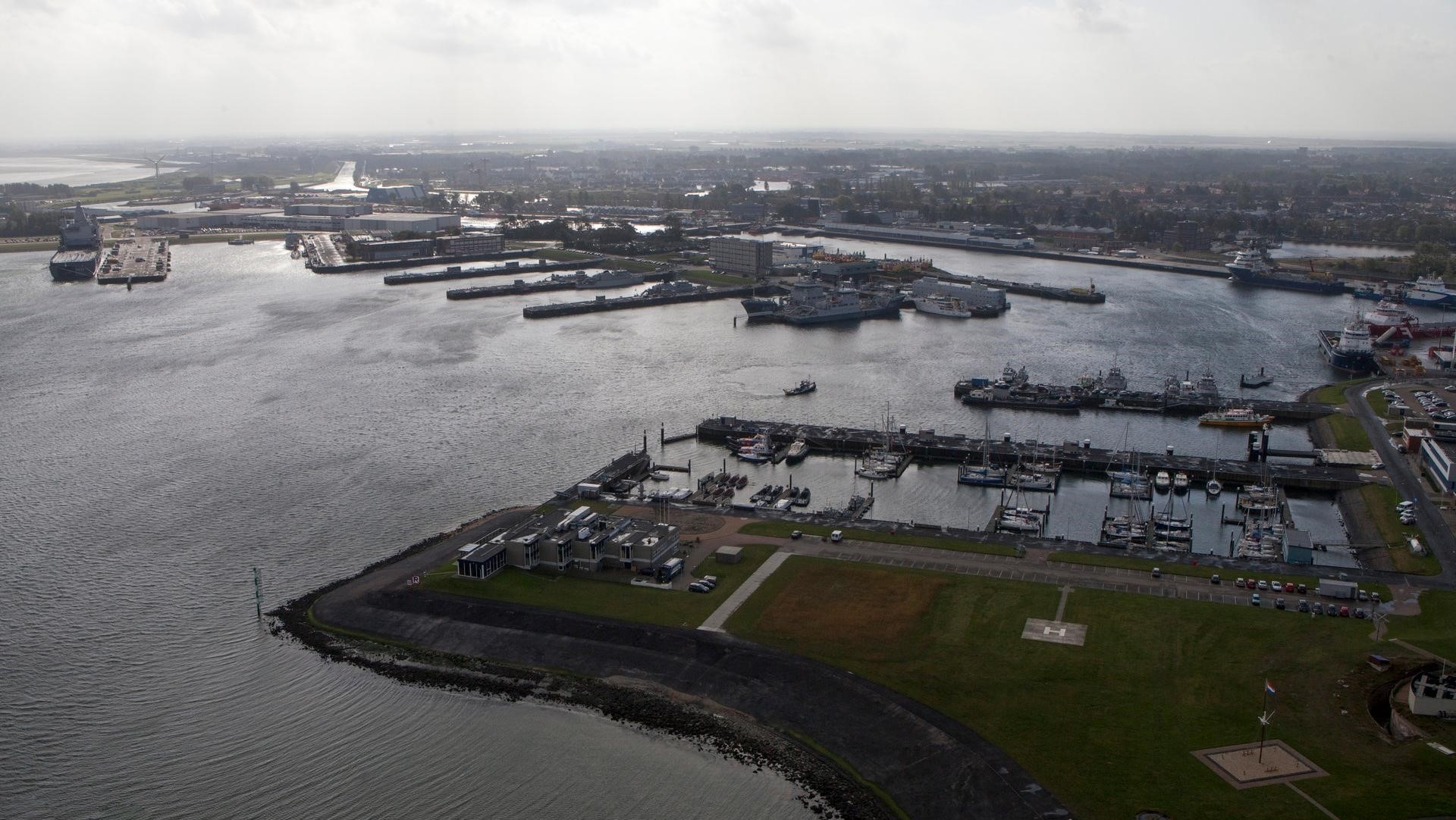 Luchtfoto van een haven met meerdere schepen aangemeerd aan kades. Op de voorgrond zijn grasvelden en enkele gebouwen te zien, terwijl de achtergrond een wijd uitzicht over het water en de stad biedt.