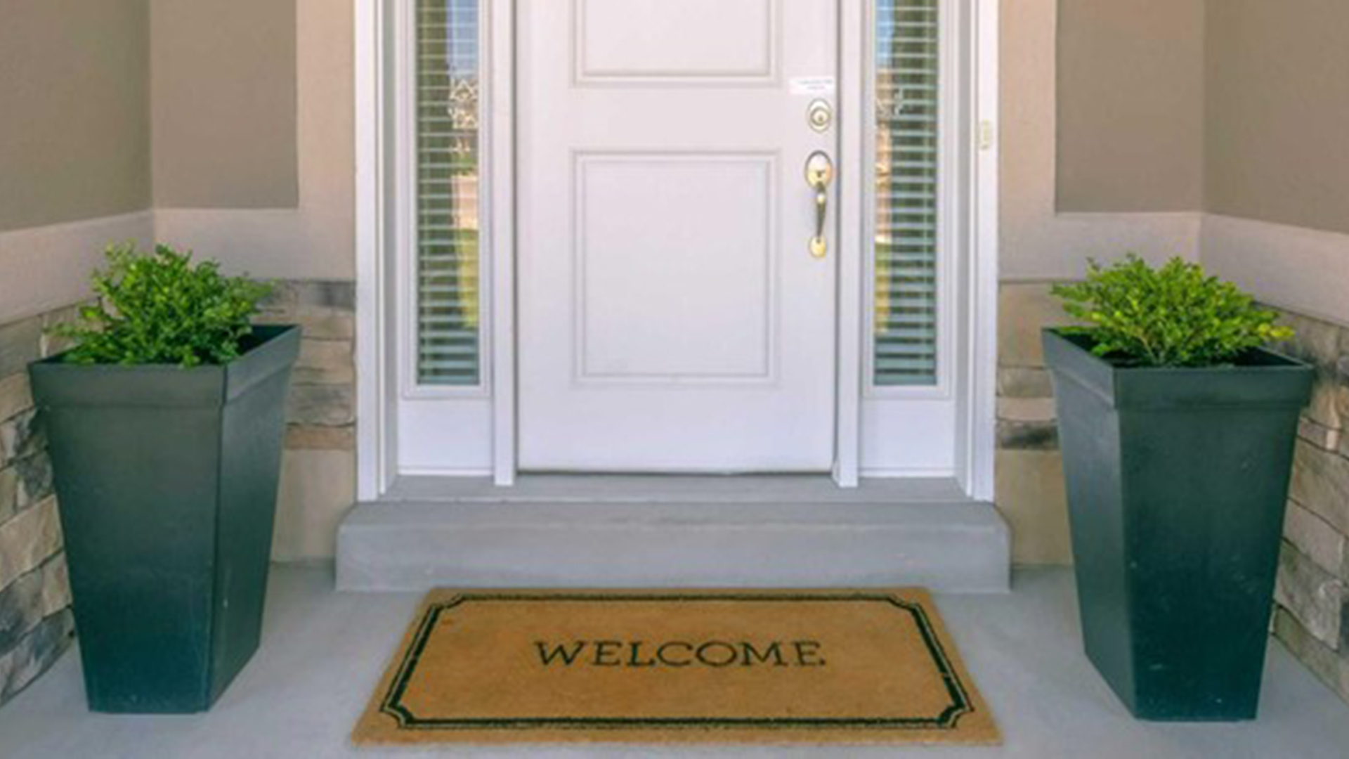 Ingang van een huis met een witte voordeur, geflankeerd door glazen panelen en twee grote potten met groene planten, en een deurmat met de tekst "Welcome".