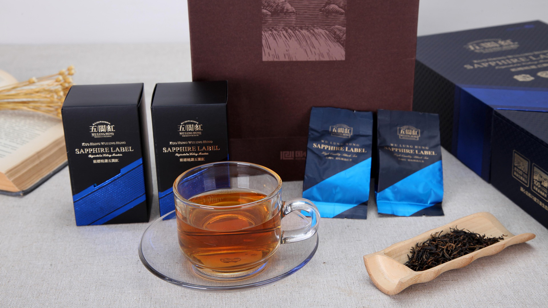 Een glas thee naast twee verpakkingen van het merk "Sapphire Label" op een tafel, met gedroogde thee in een houten schaal en een open boek op de achtergrond.