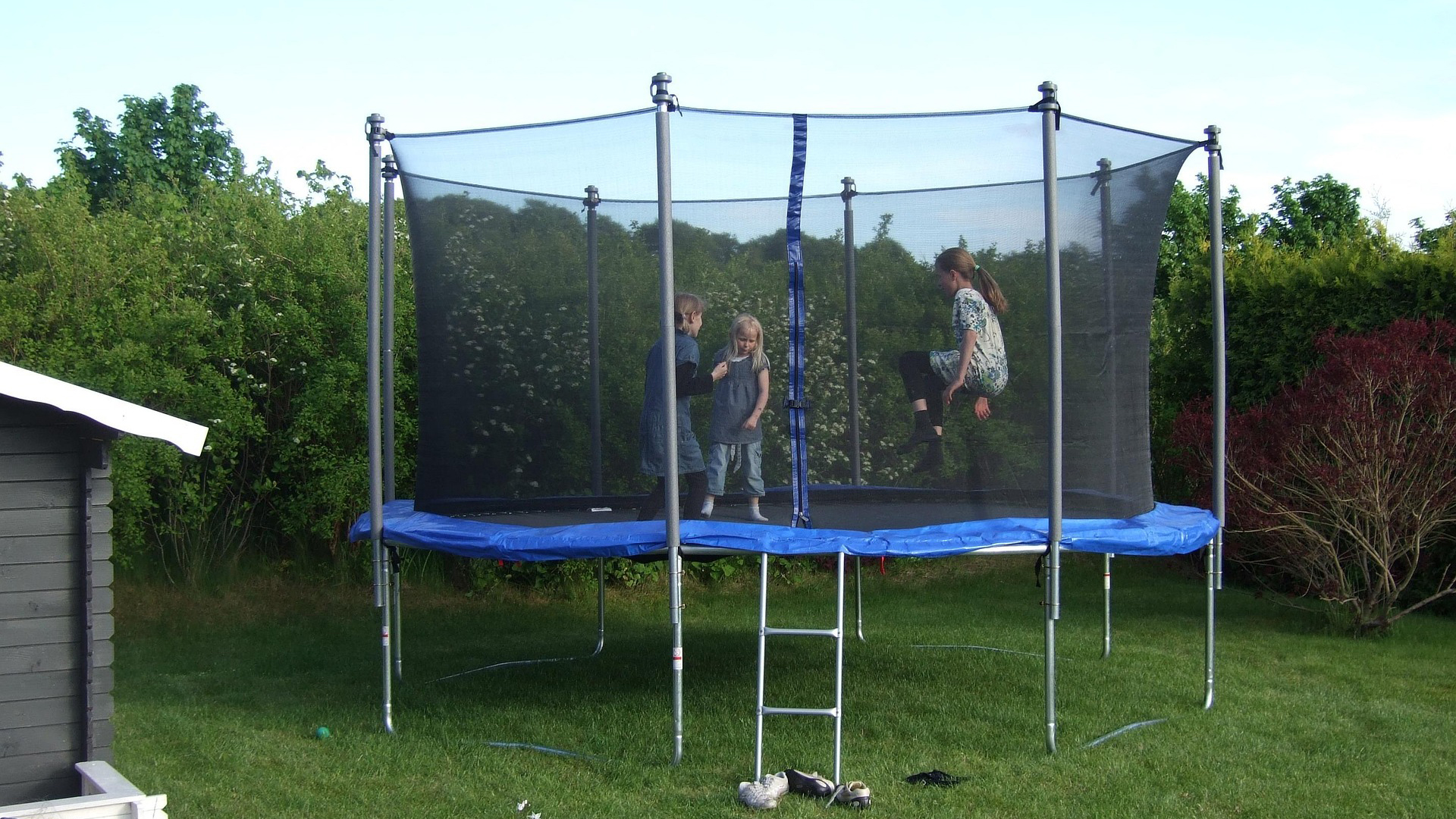 Drie kinderen spelen op een trampoline met een veiligheidsnet in een groene tuin.