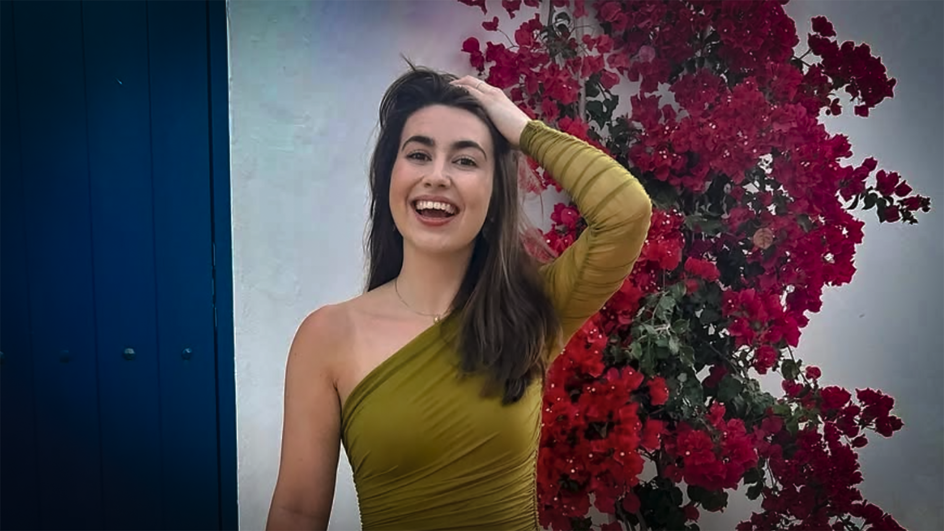 Vrouw in een olijfgroene jurk met één schouder poseert glimlachend voor een achtergrond van rode bougainvillea bloemen en een blauwe deur.