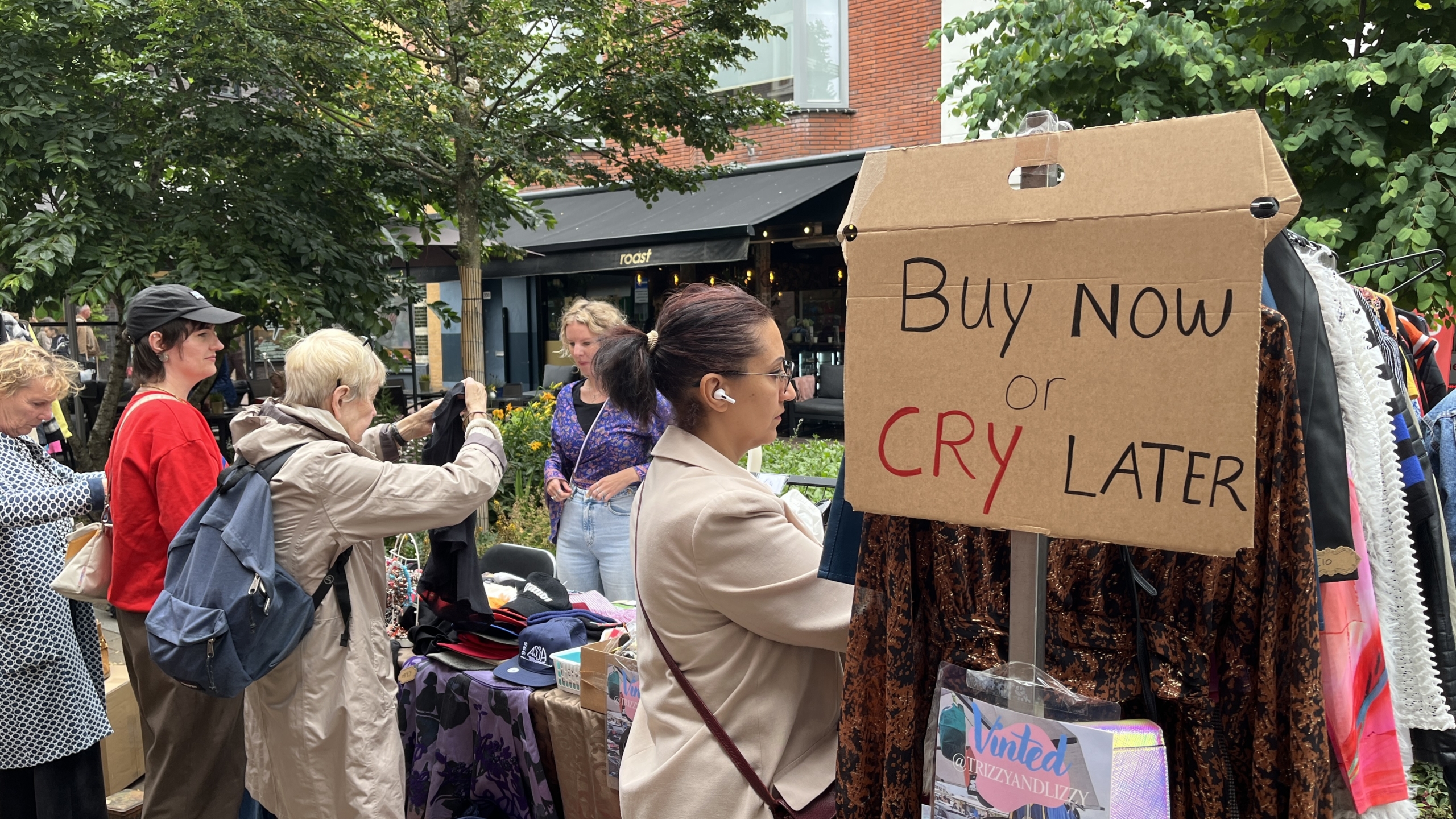 Mensen bekijken kleding bij een kraam op een openluchtmarkt met een kartonnen bord waarop "Buy Now or Cry Later" staat.