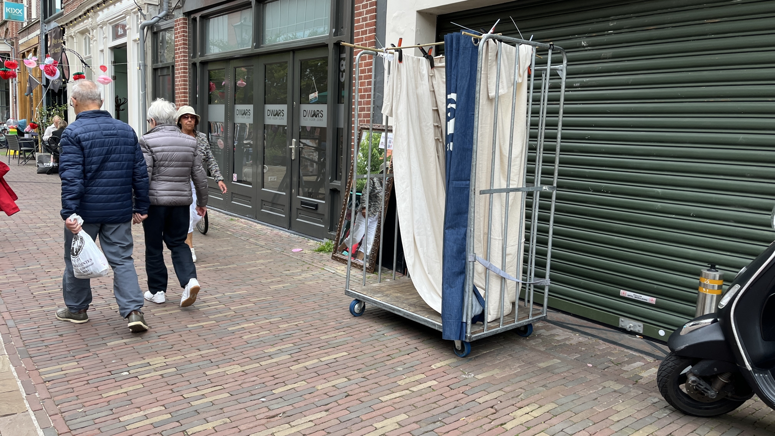 Twee mensen lopen op een stenige straat langs een winkel met een gesloten rolluik. Er staat een rek met textiel en een spiegel tegen de muur. Rechts is een scooter te zien.