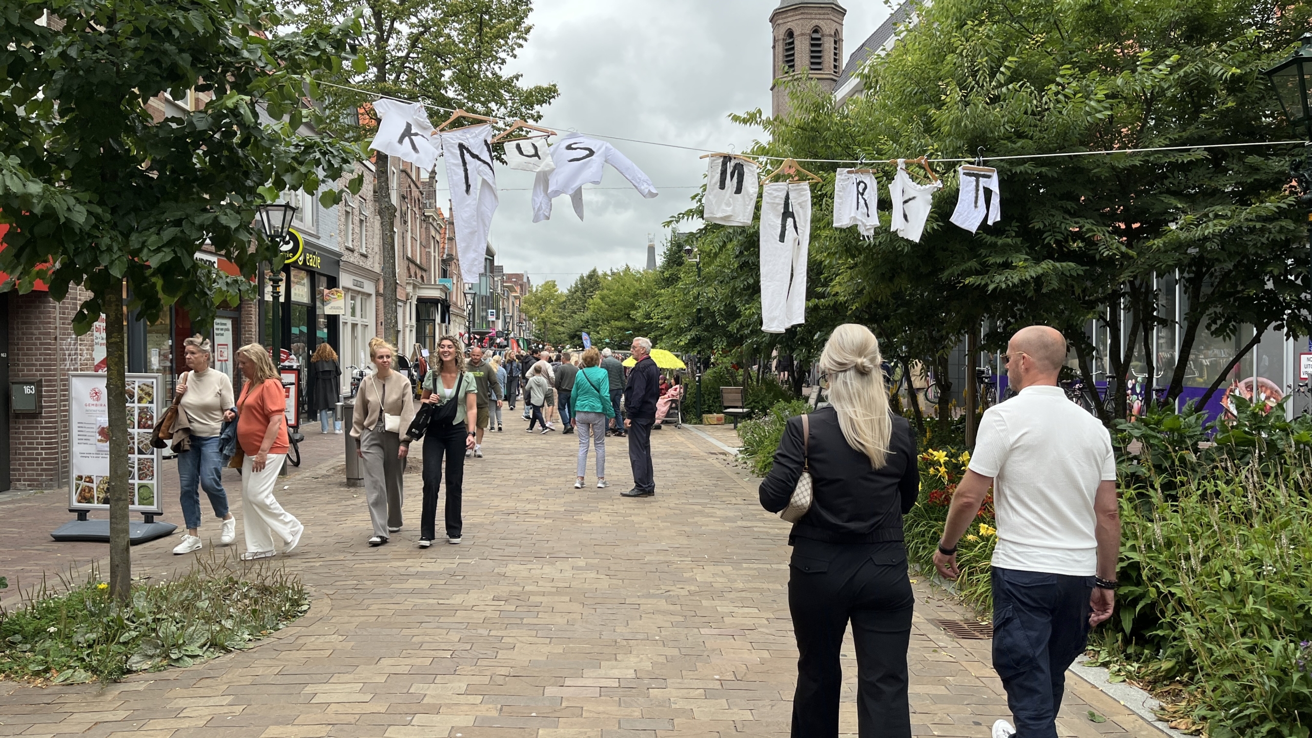 Een drukke straatmarkt met mensen die langs winkels en kraampjes lopen, met een banner van witte kledingstukken met de woorden 'Kunst Markt' erboven.