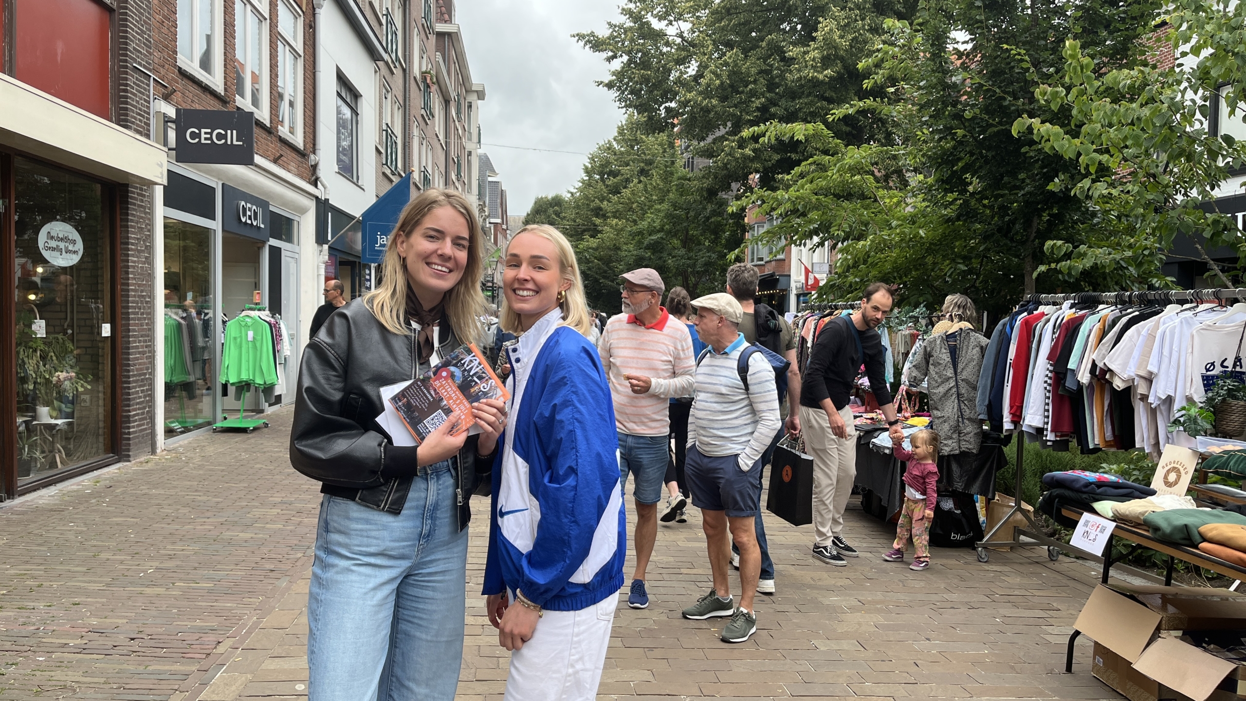 Twee vrouwen poseren glimlachend voor de camera op een drukke winkelstraat, terwijl ze flyers vasthouden; op de achtergrond staan mensen te winkelen bij kledingrekken langs de straat.