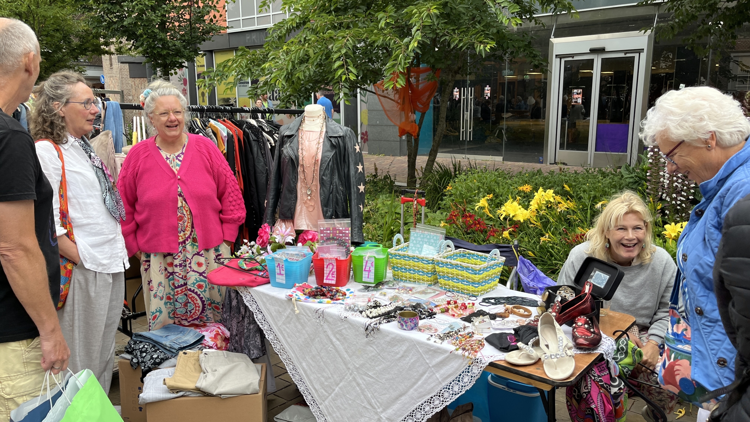 Mensen staan bij een kraam op een rommelmarkt met kleding, sieraden en andere goederen uitgestald op tafels. Er is een mannequin met een jas en ketting te zien. Enkele personen lachen en praten met elkaar, terwijl anderen de spullen bekijken. Op de achtergrond zijn winkels en beplanting zichtbaar.