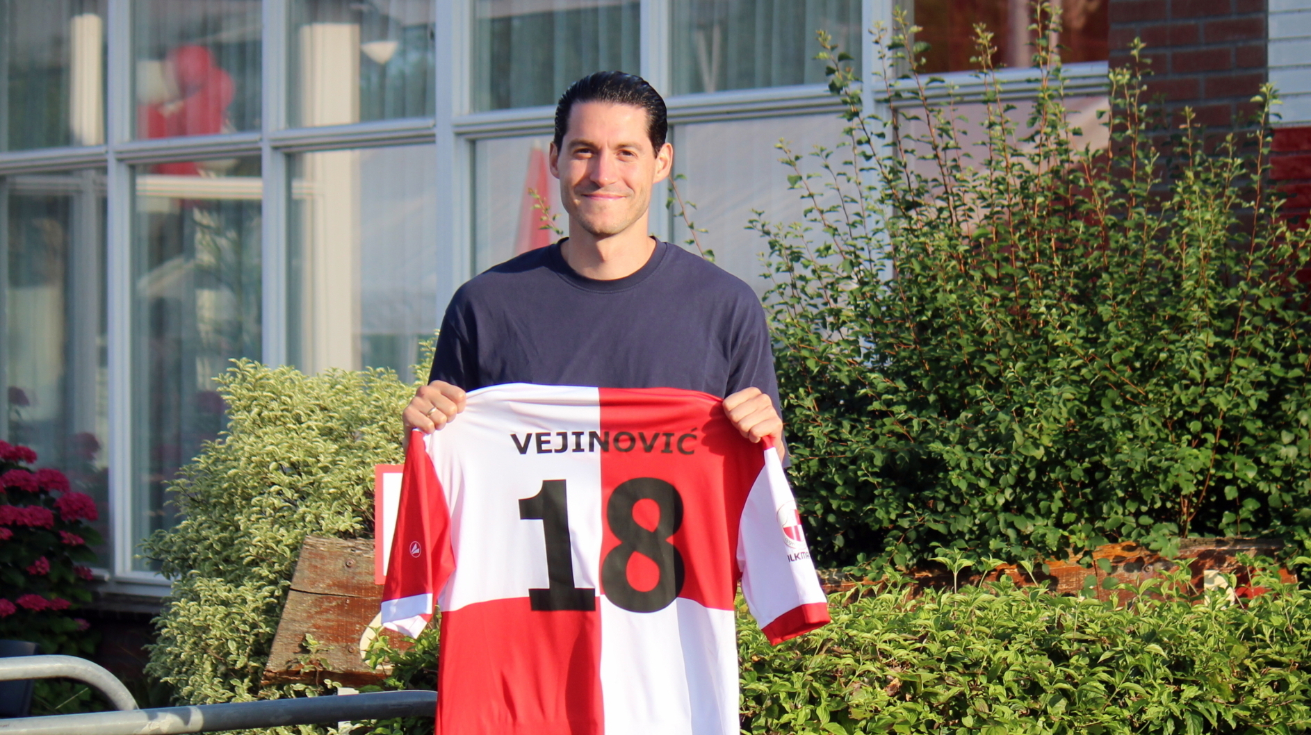 Een persoon houdt een rood-wit voetbalshirt vast met de naam 'Vejinović' en nummer 18, staand voor een gebouw met planten op de voorgrond.