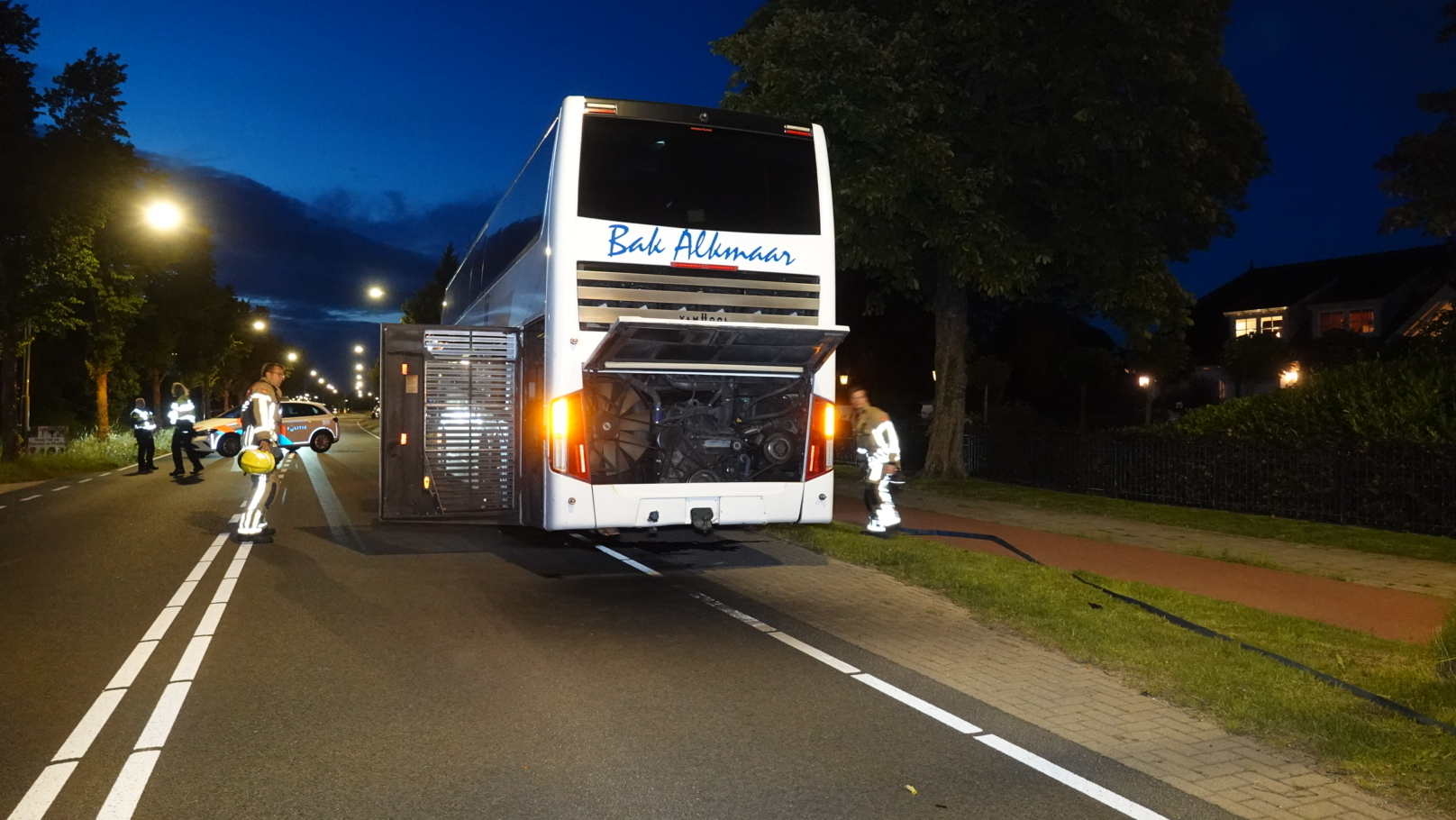 Brandweerlieden werken 's nachts aan de achterkant van een stilstaande dubbeldekkerbus met een geopende motorkap, terwijl een politiewagen de weg blokkeert.