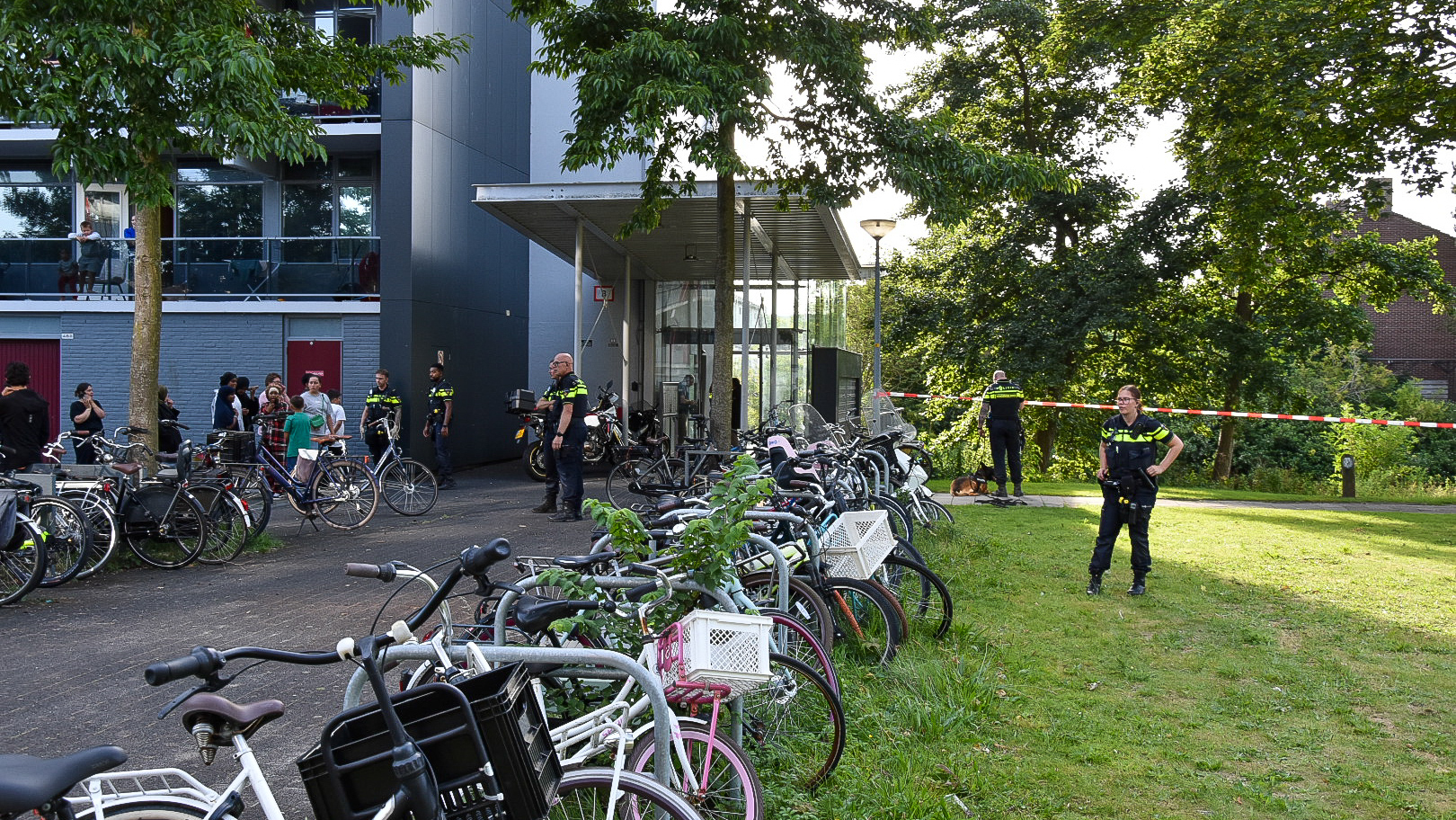 Politieagenten bij een flatgebouw met fietsenrekken ernaast, afgezet met rood-wit politielint, terwijl mensen toekijken.