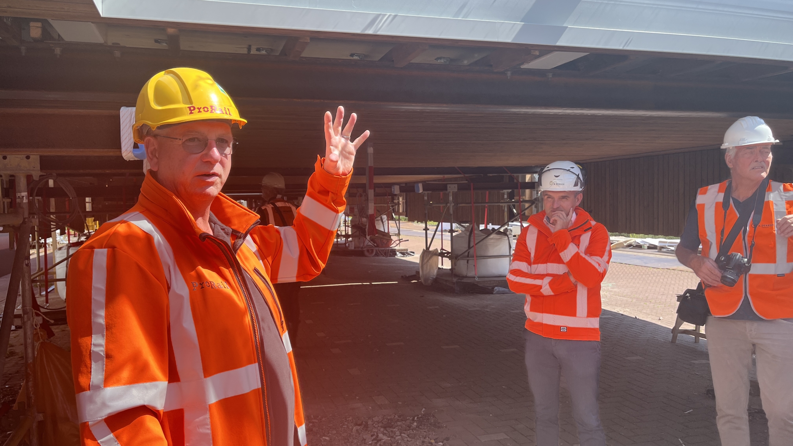 Drie mannen in oranje veiligheidsjassen en helmen staan bij een bouwplaats, waarvan één man iets uitlegt met een opgestoken hand.