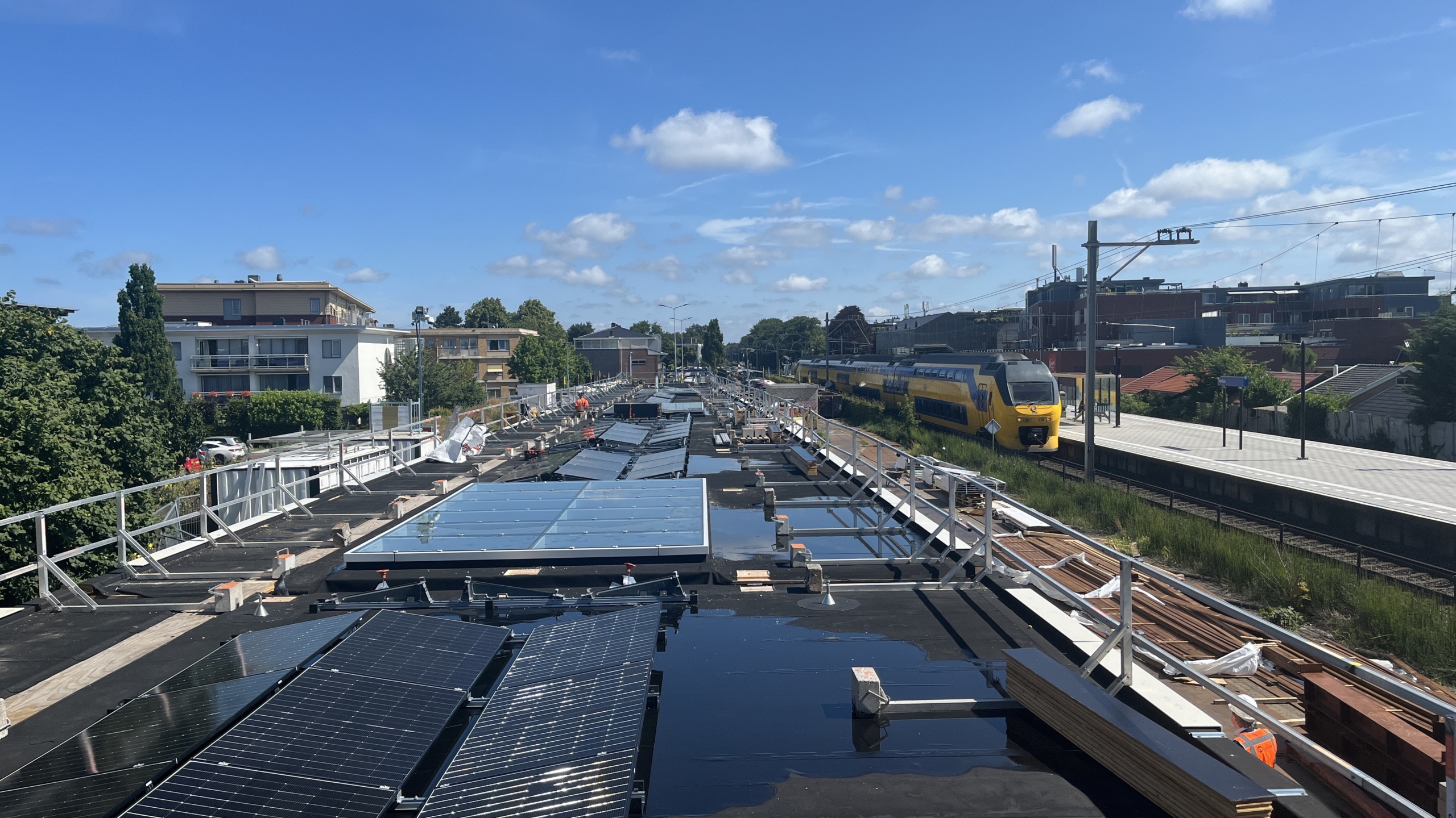Zonnig treinstation met zonnepanelen op het dak en een gele passagierstrein op het spoor, omgeven door gebouwen en groene bomen.