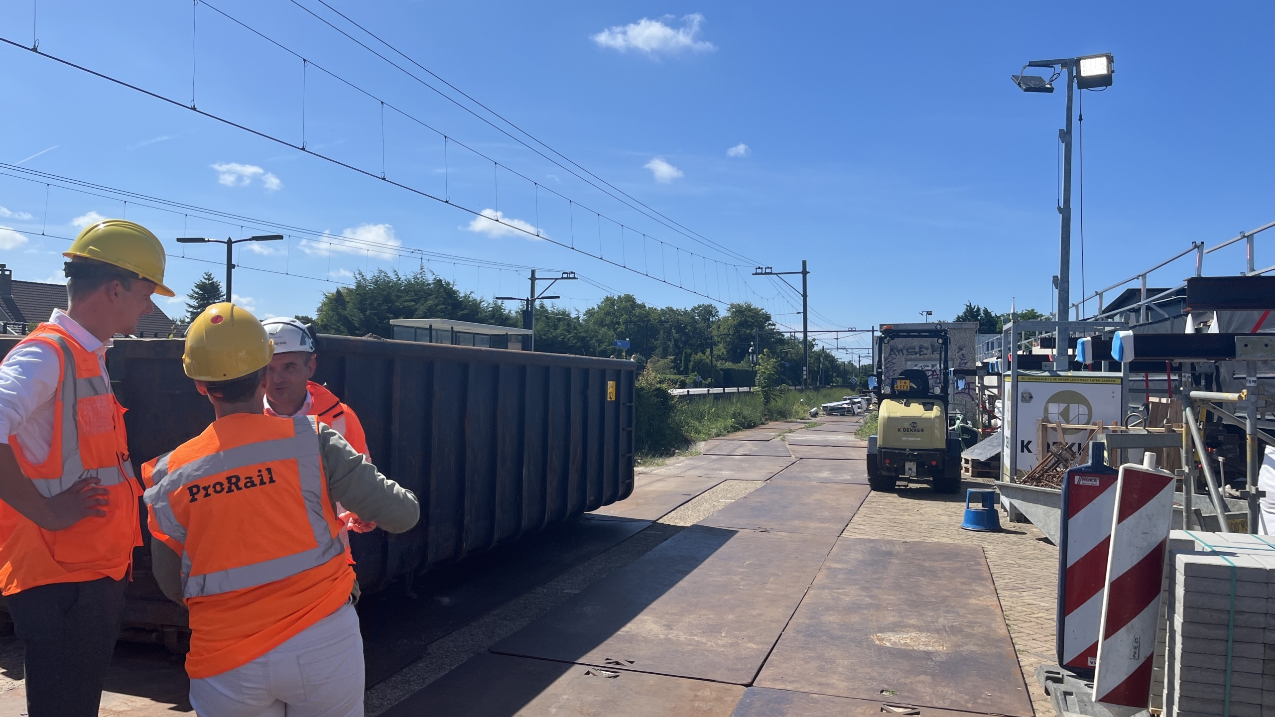 Mensen met helmen en ProRail-veiligheidsvesten staan te praten bij een bouwlocatie naast een spoorlijn, met bouwmateriaal en apparatuur in de achtergrond, op een zonnige dag.