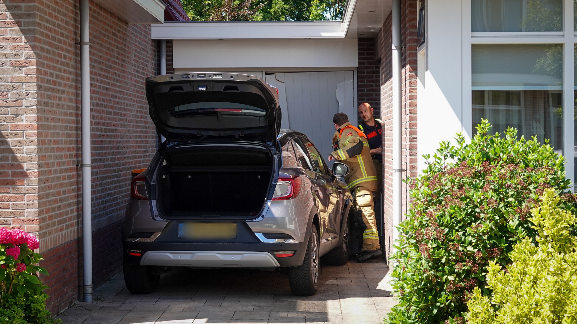 Twee brandweermannen onderzoeken een auto die tegen een muur van een huis is gebotst in een smalle oprit, met de achterklep open.