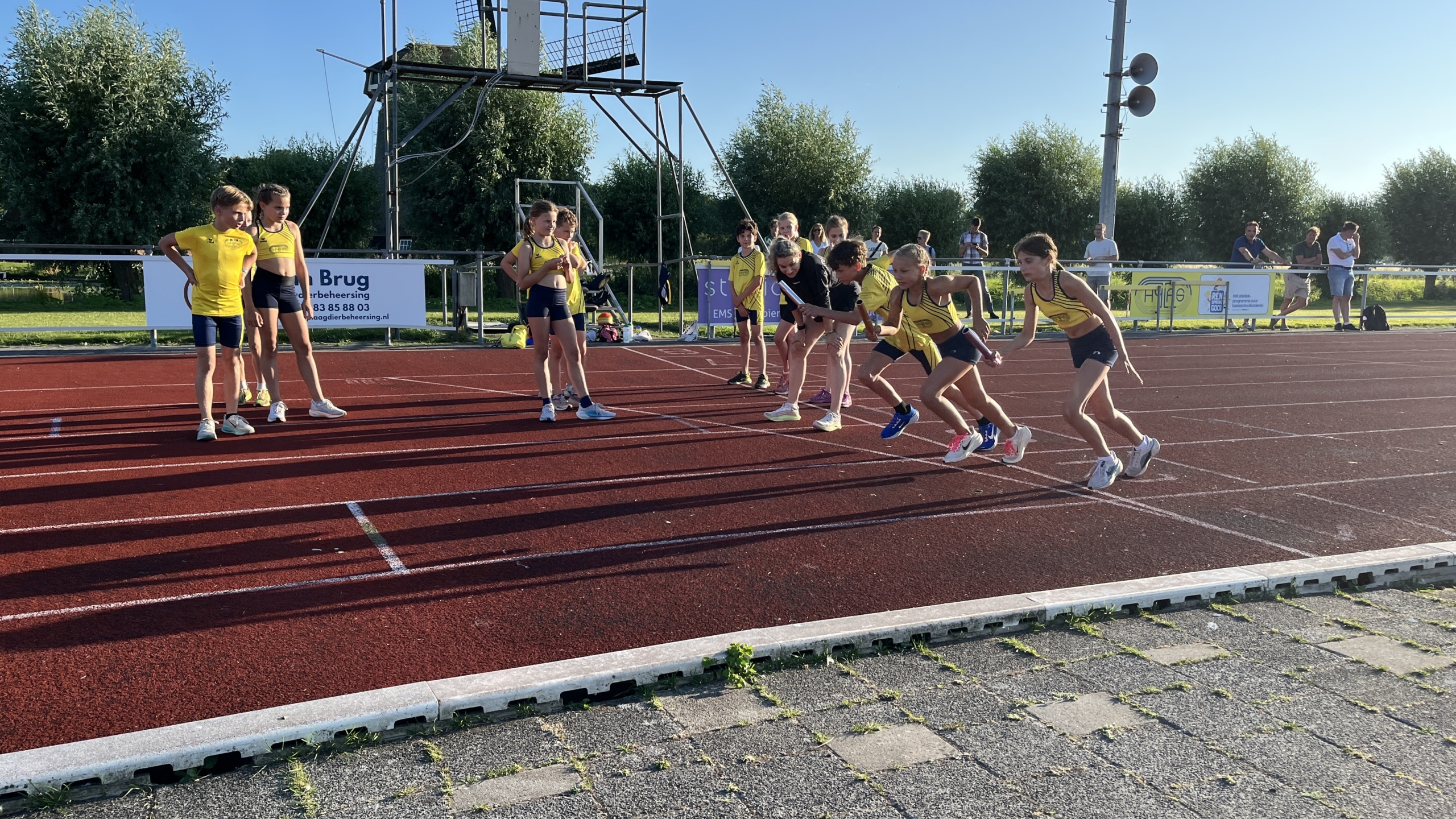 Kinderen in sportkleding staan klaar om te rennen op een atletiekbaan op een zonnige dag, met toeschouwers in de achtergrond.