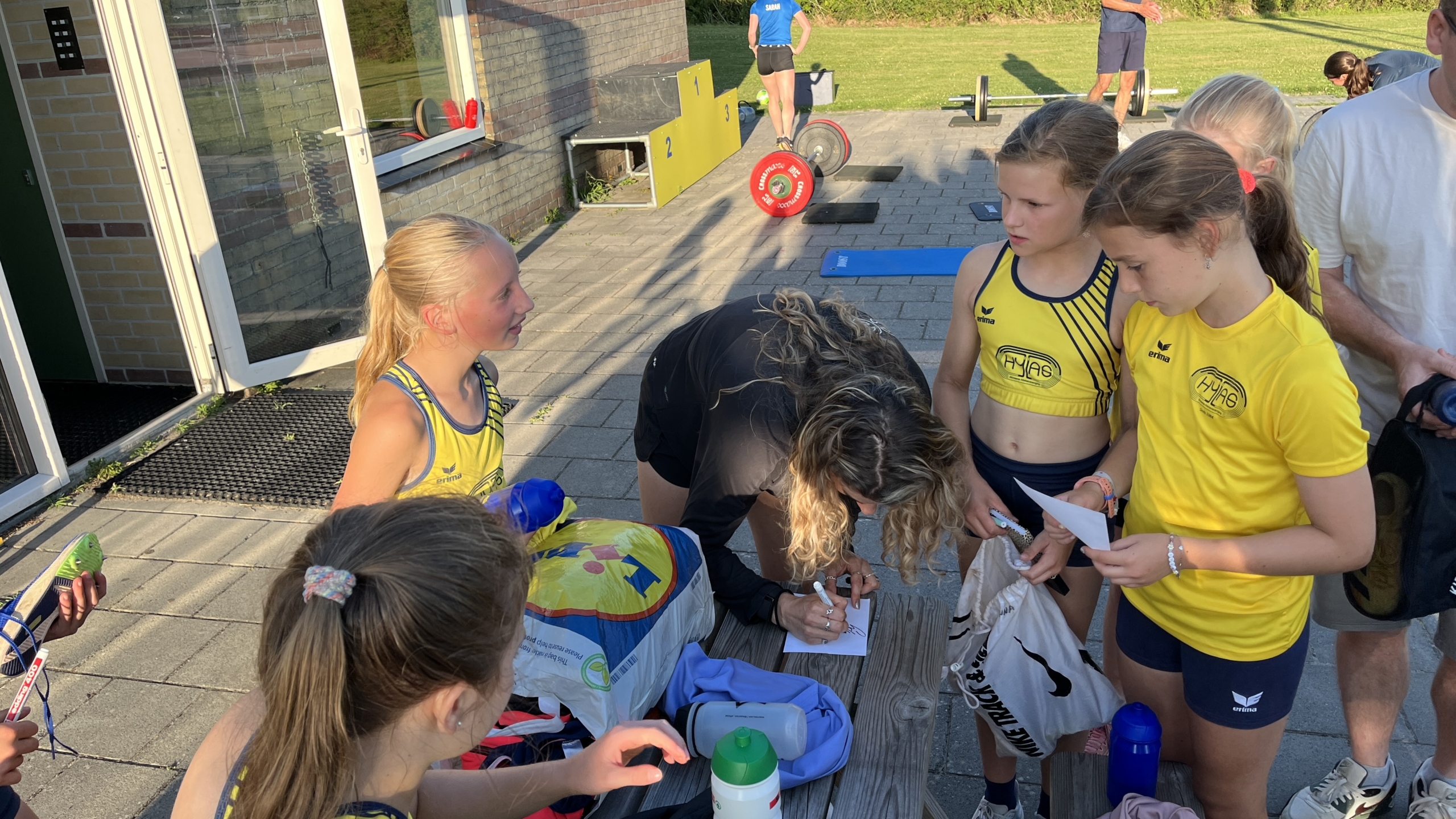 Jonge atletes in sportkleding verzamelen zich rond een vrouw die iets op een papier schrijft, met sportuitrusting op de achtergrond.