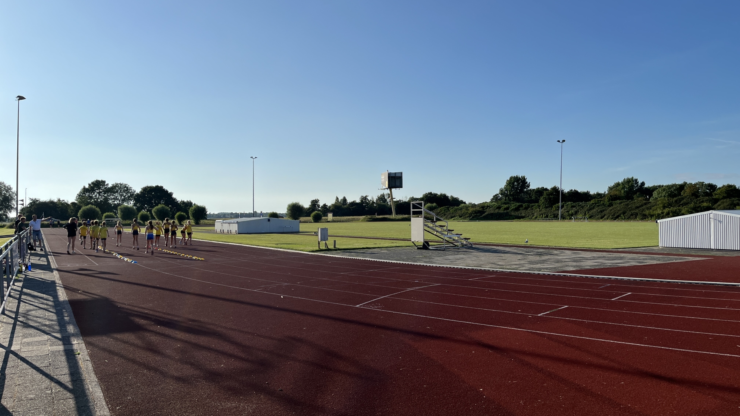 Atletiekbaan met sporters in gele kleding, begeleid door volwassenen, onder een heldere blauwe hemel.