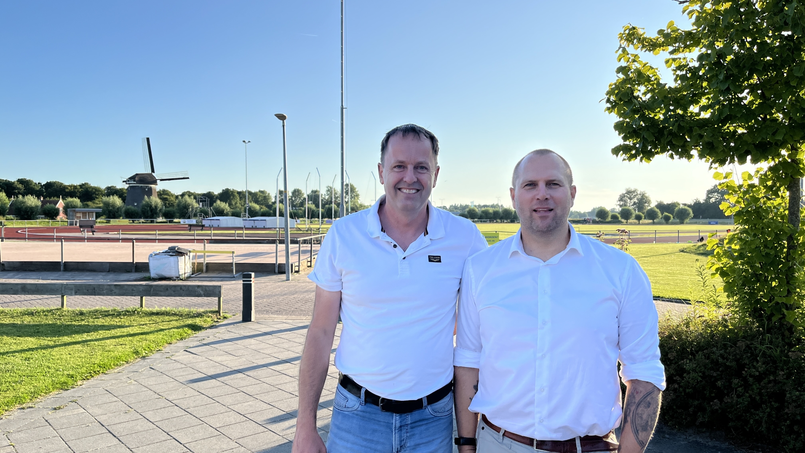 Twee mannen staan buiten op een zonnige dag voor een atletiekbaan met een traditionele Nederlandse windmolen op de achtergrond.