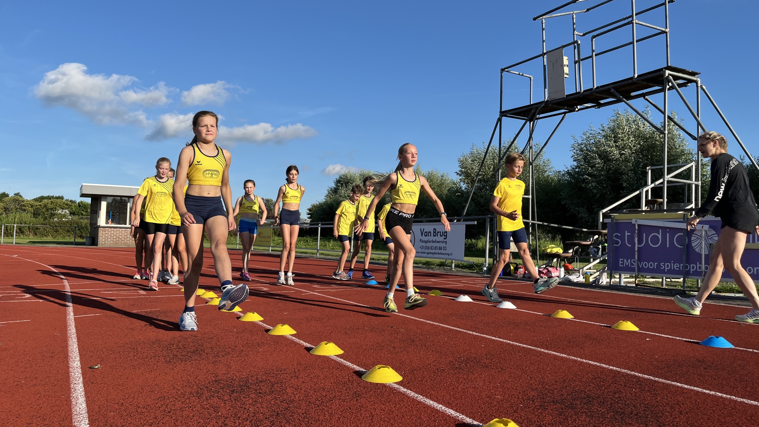 Jonge atleten in gele sportkleding trainen op een atletiekbaan met kegels onder een heldere blauwe lucht.