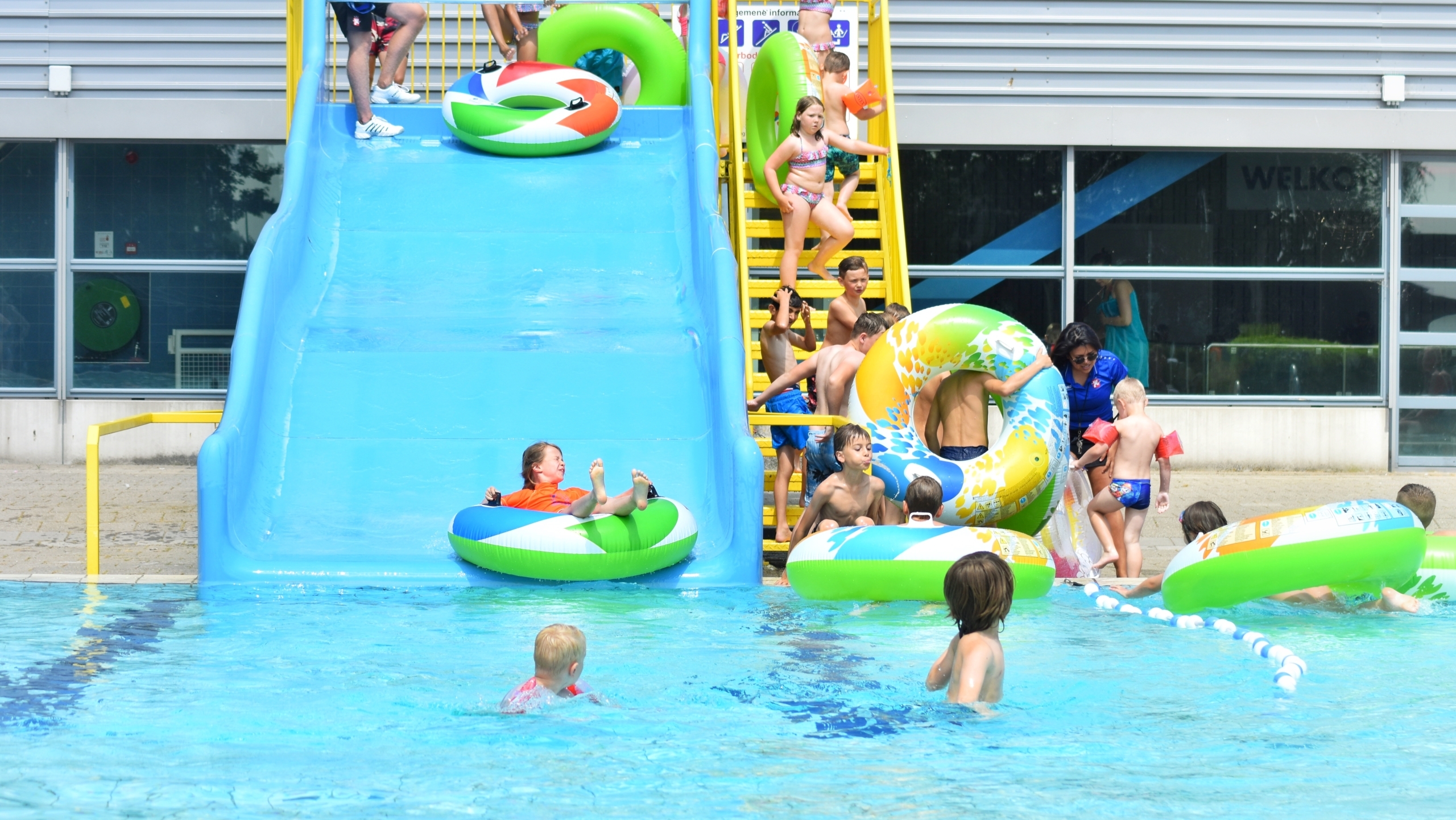 Kinderen spelen in een zwembad met een blauwe glijbaan, sommige met grote groene en kleurrijke zwemringen.