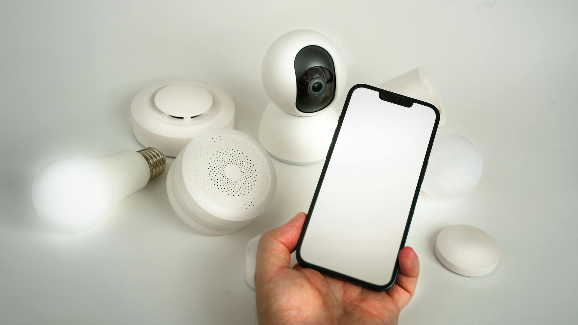 Een hand houdt een smartphone vast met een leeg scherm, omringd door verschillende slimme apparaten, waaronder een gloeilamp, een camera en een luidspreker.